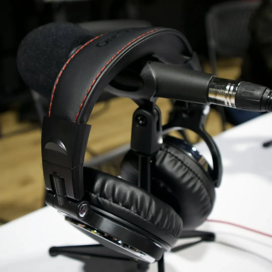 Black over-ear headphones with padded ear cups and headband, placed on a stand with a microphone attached, on a white surface.