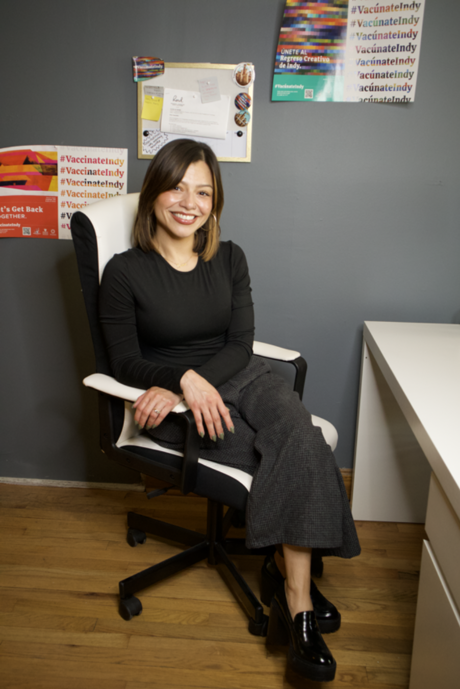 A woman sitting on an office chair in a room with a gray wall, smiling at the camera. There are posters and a bulletin board on the wall behind her related to vaccination awareness.
