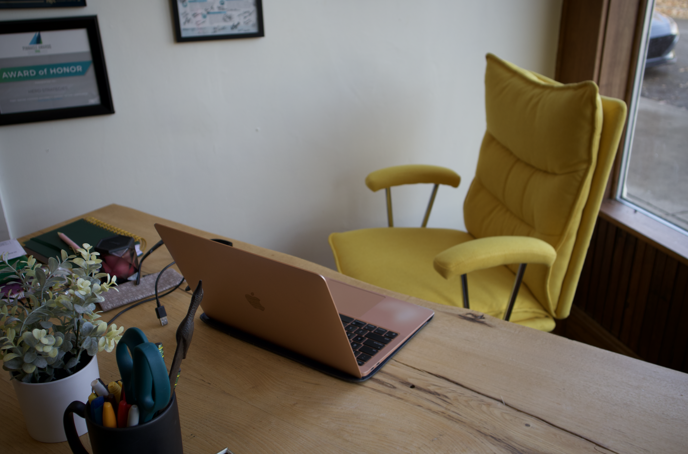 A wooden desk with a pink MacBook, a potted plant, scissors, and pens. In the background, a yellow armchair near a window, with framed certificates or artwork on the wall.
