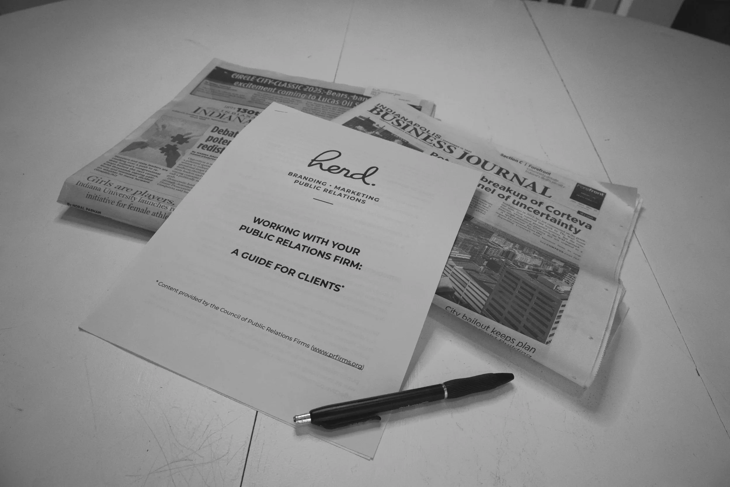 A printed guide titled 'Working with Your Public Relations Firm: A Guide for Clients' lies on top of a stack of newspapers on a white table. A black pen is placed next to the guide.