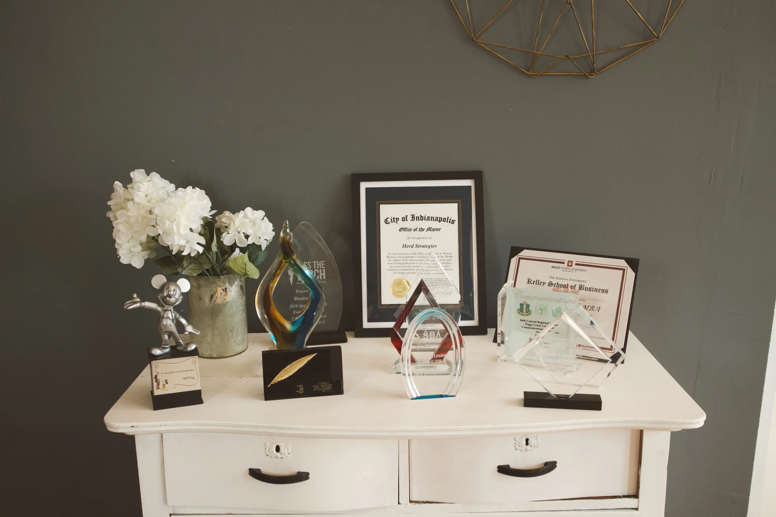 Awards, certificates, and plaques displayed on a white dresser, with a potted white hydrangea plant and a silver Mickey Mouse figurine. The background wall is painted dark green and features a geometric wire wall decor.