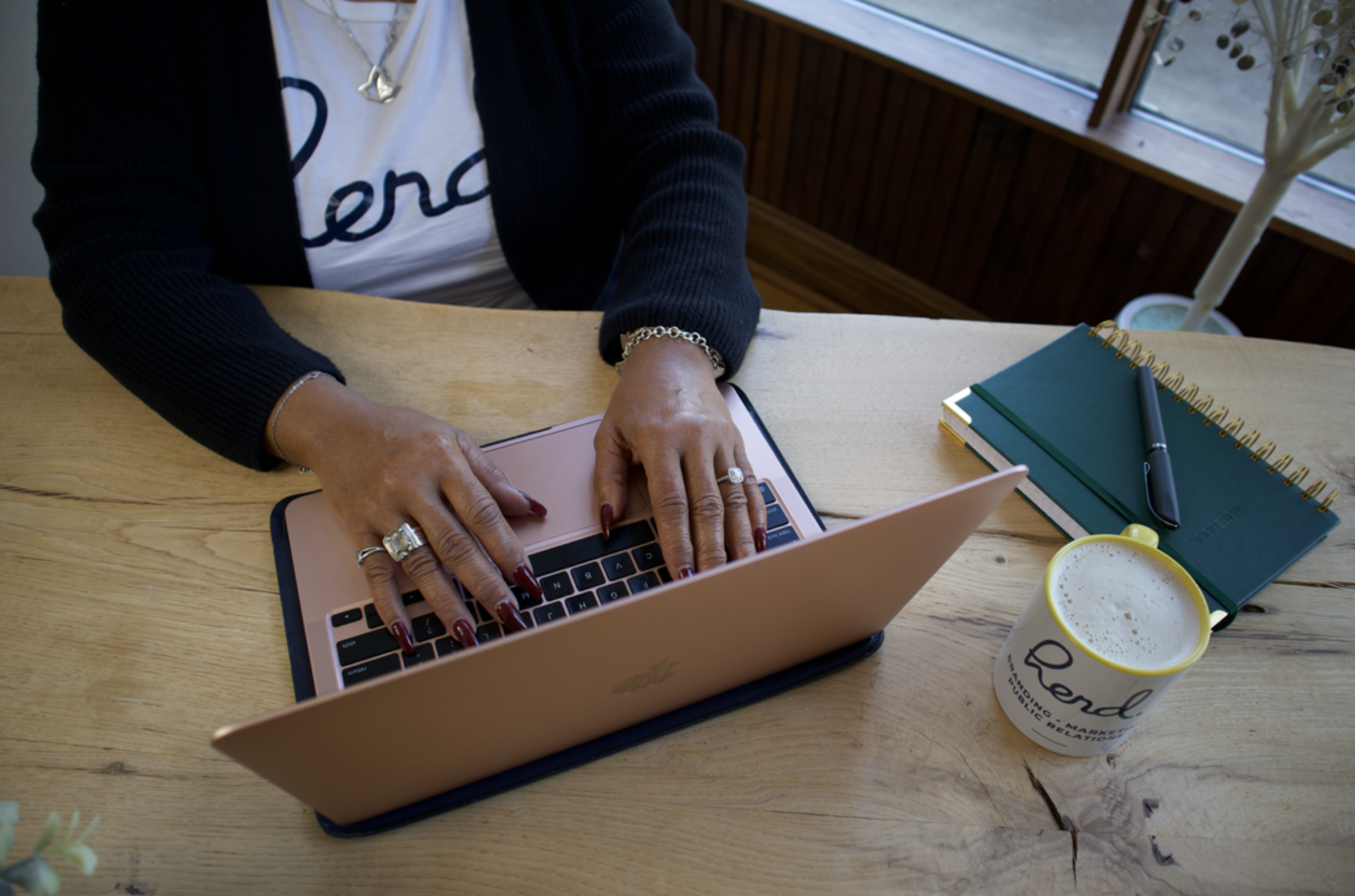 Person working on a MacBook at a wooden desk, wearing jewelry, with notebooks, pen, and a cup of coffee on the desk.