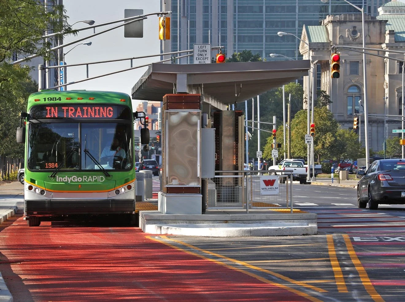 A green and white IndyGo Rapid bus at a bus stop on a city street, with a digital display reading "IN TRAINING," red lights on traffic signals, and tall buildings in the background.