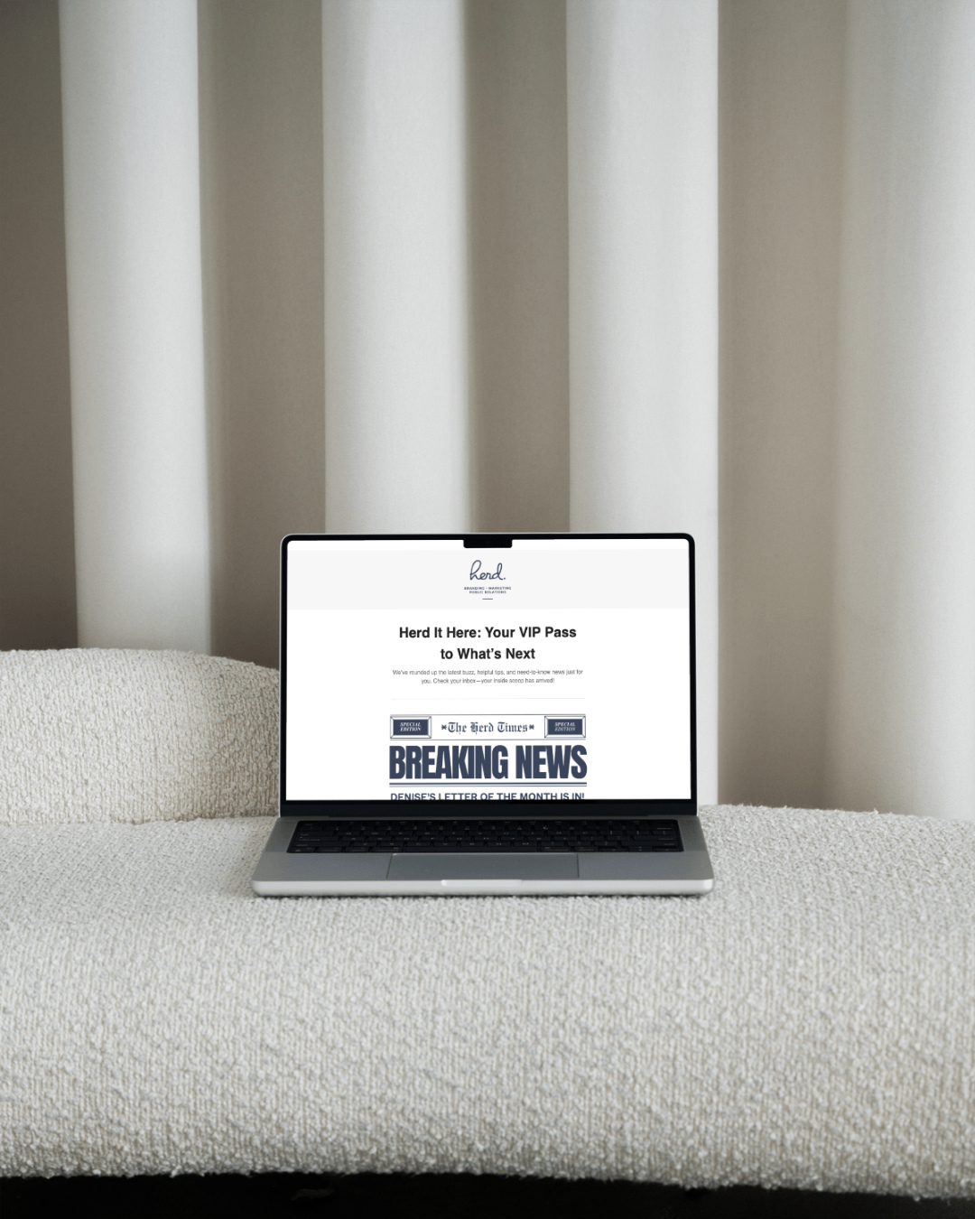 Laptop on a beige textured surface displaying a newsletter titled 'Herd It Here: Your VIP Pass to What's Next' with a headline 'BREAKING NEWS' and a white background, against a background of light-colored curtains.