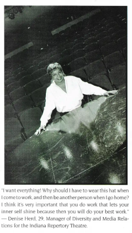 A woman, leaning on a theater or auditorium stage with empty seats in the background, smiling at the camera.