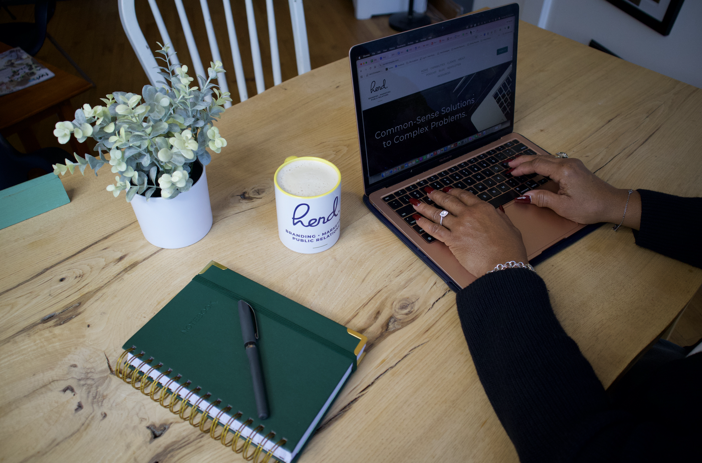 Person typing on a laptop at a wooden table with a potted plant, a green notebook with a pen, and a cup of coffee.