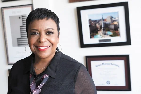 Portrait of a woman with short hair, wearing a black blouse and a striped tie, smiling in an office with framed pictures and a certificate on the wall behind her.