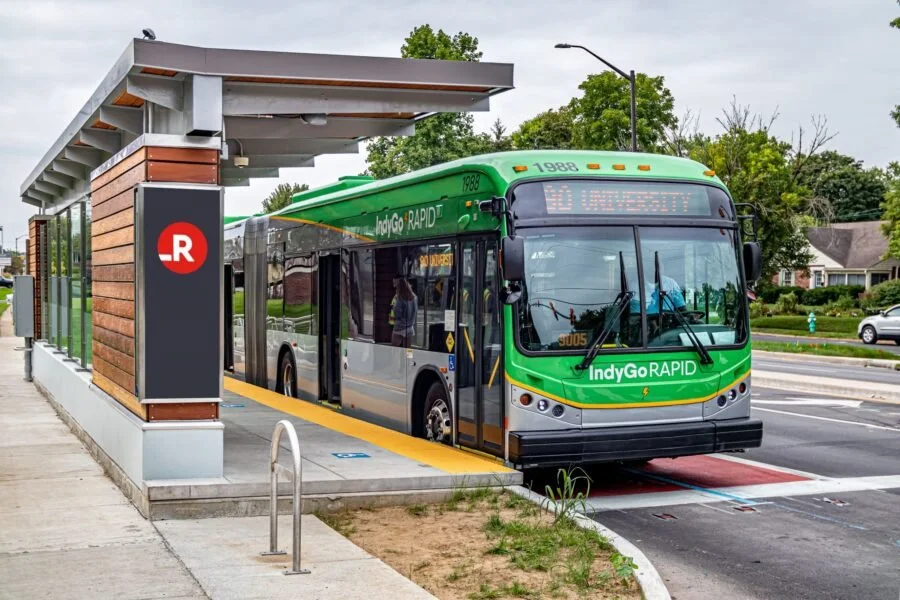 A green and gray IndyGo RAPID bus at an outdoor bus station in a suburban area with trees and houses in the background.