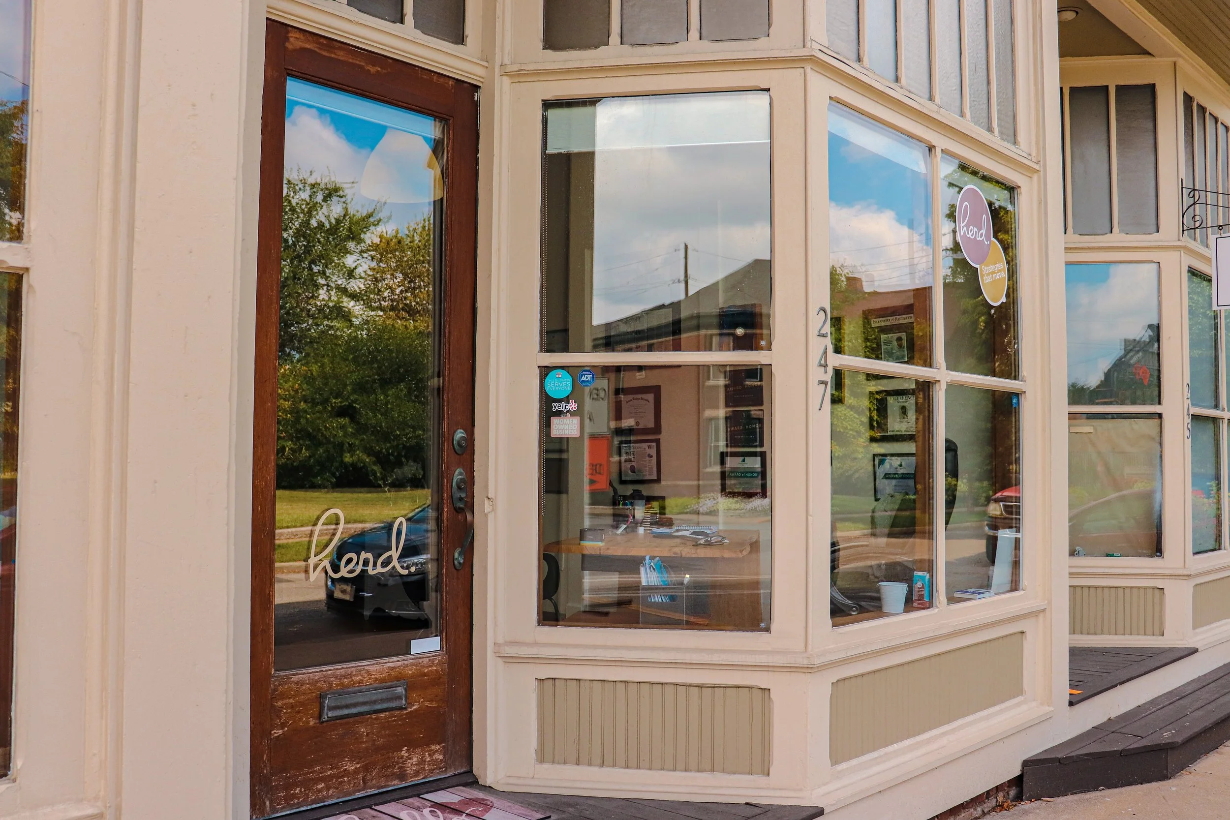 Exterior view of a storefront with a wooden door that has a glass panel and the word "hend" written on it, and large glass windows displaying interior office space, building number 247, and a sidewalk in front.
