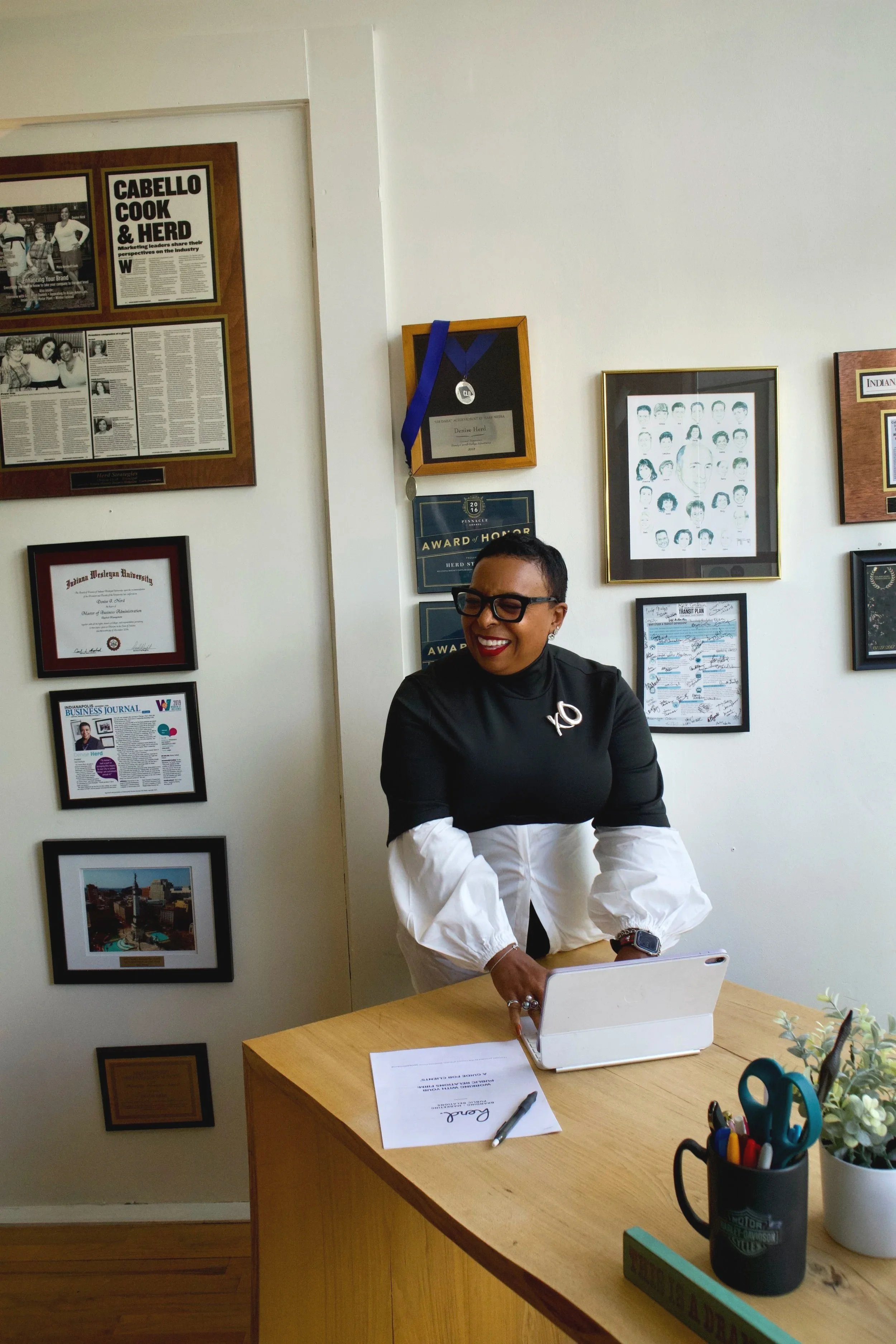 A woman with short dark hair and glasses, wearing a black top with white long sleeves, smiling and working on a laptop at a wooden desk in an office decorated with awards, certificates, and framed photos.