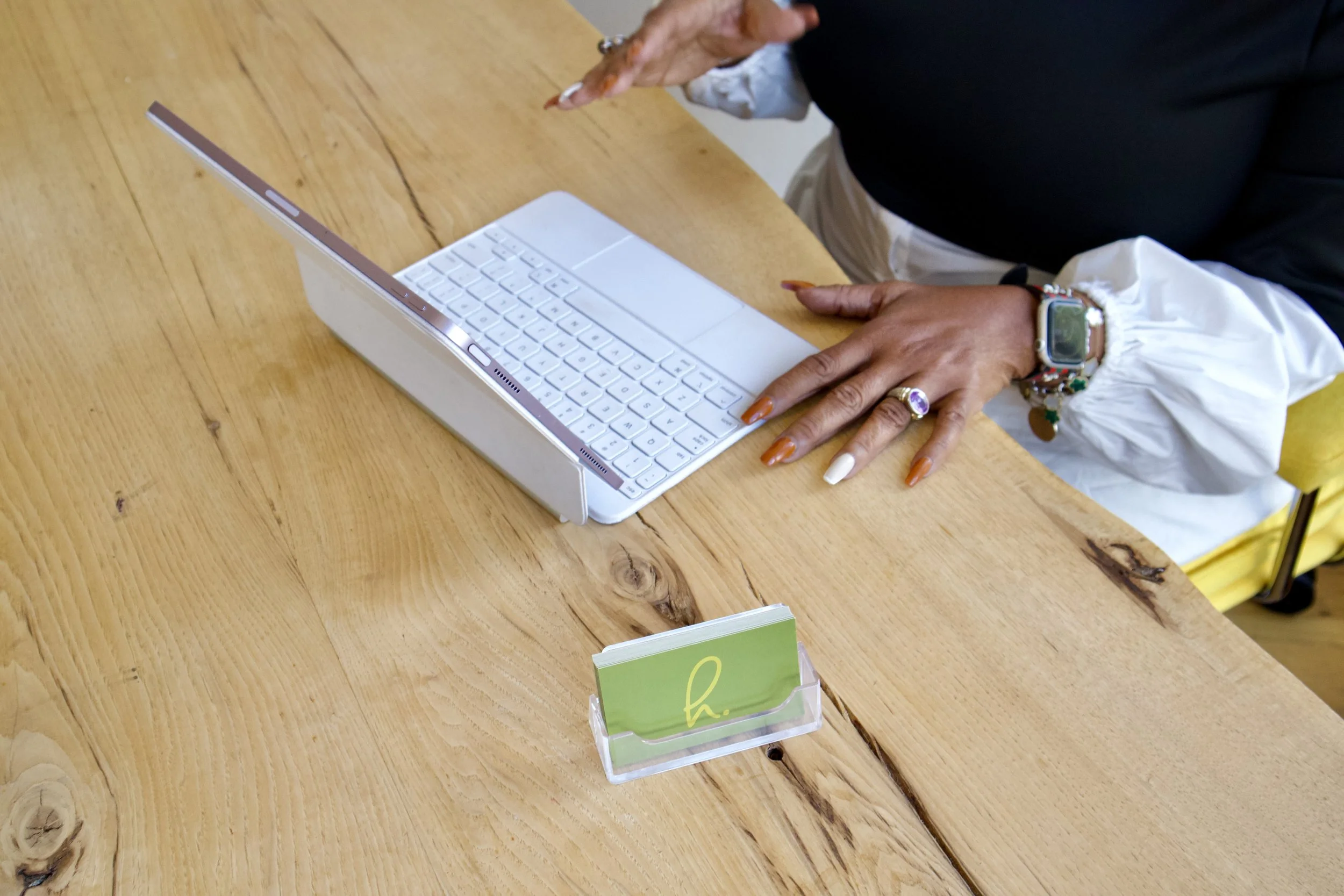Person using a white tablet with a detachable keyboard on a wooden table, with a small green card holder labeled 'h' in front.