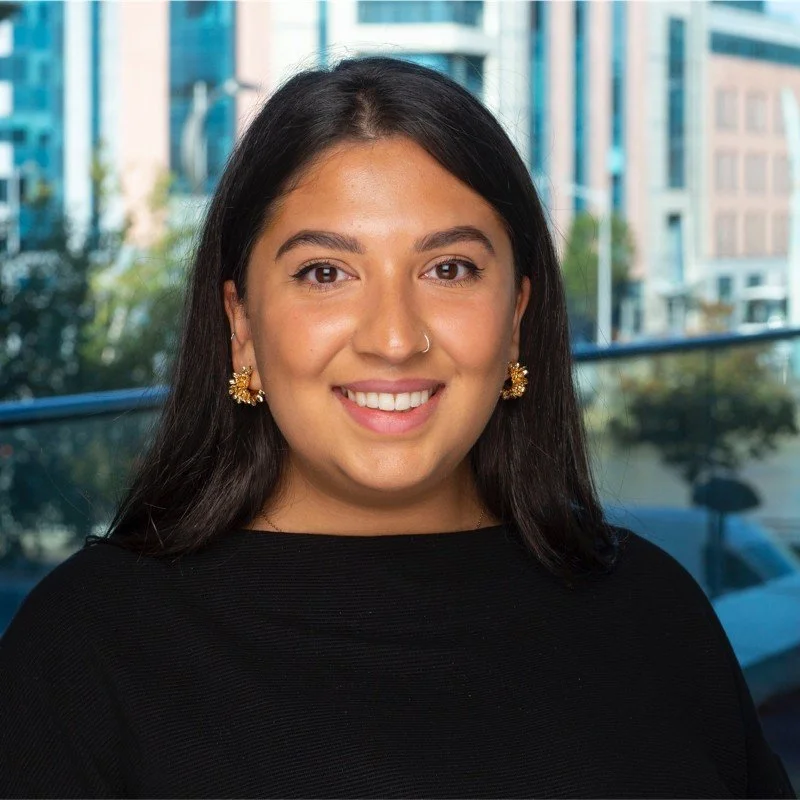 Portrait of a woman with dark hair, wearing gold earrings and a black top, smiling, with city buildings and trees in the background.