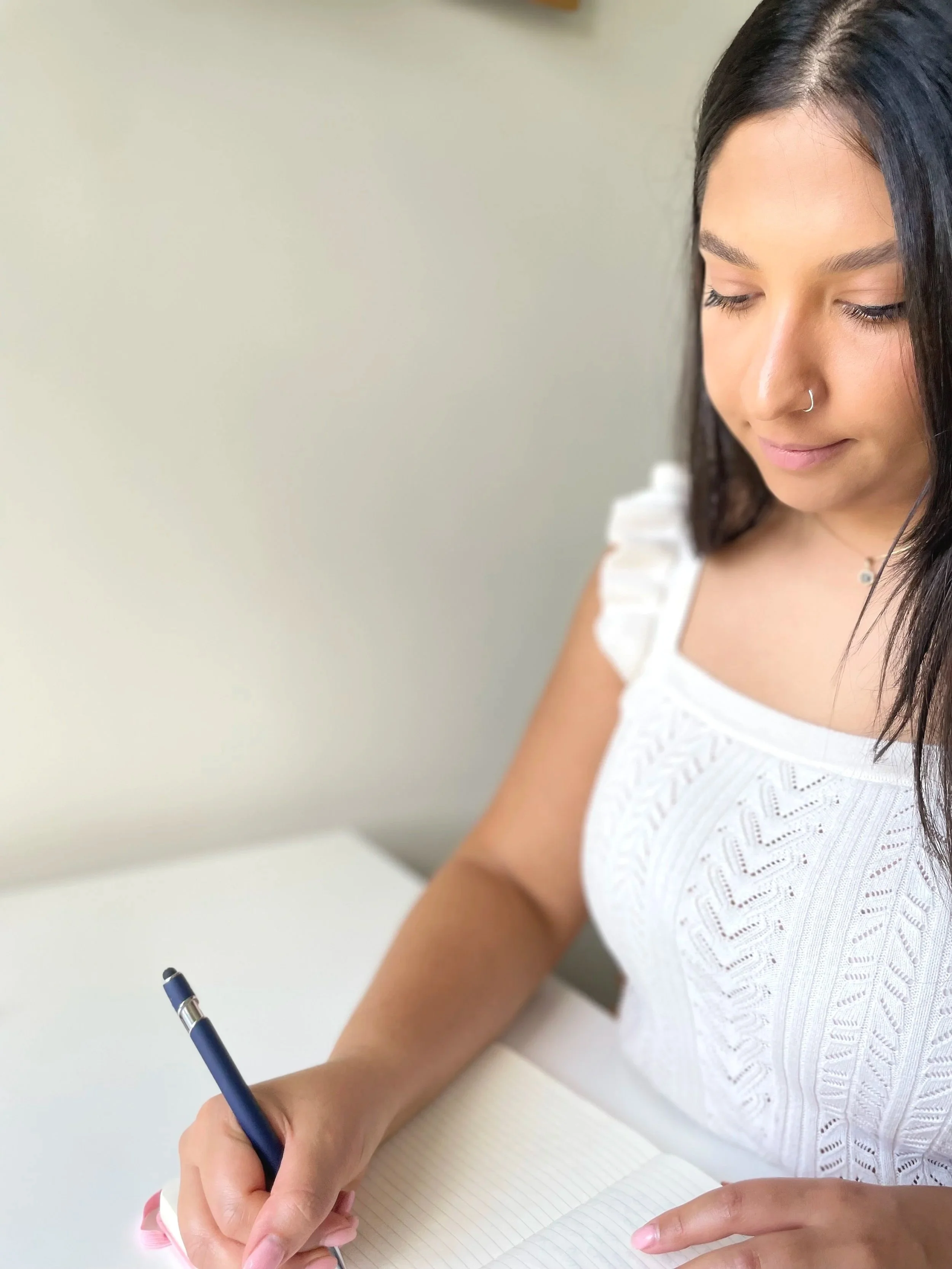 A woman with dark hair and a nose ring writing in a lined notebook with a pen.