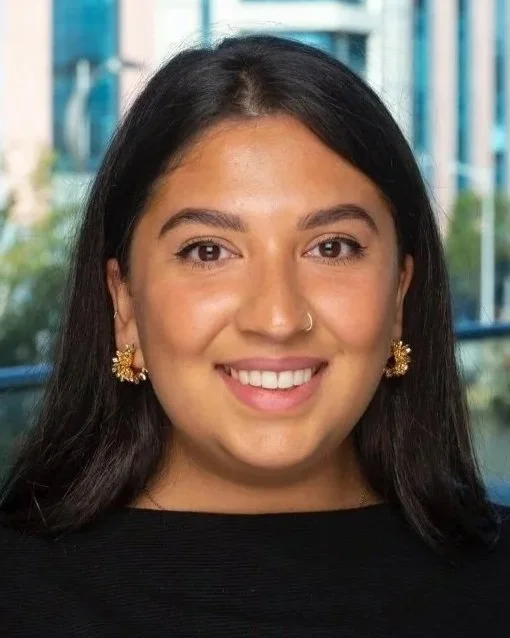 A young woman with long dark hair, wearing gold flower-shaped earrings and a nose ring, smiling in front of a cityscape window.