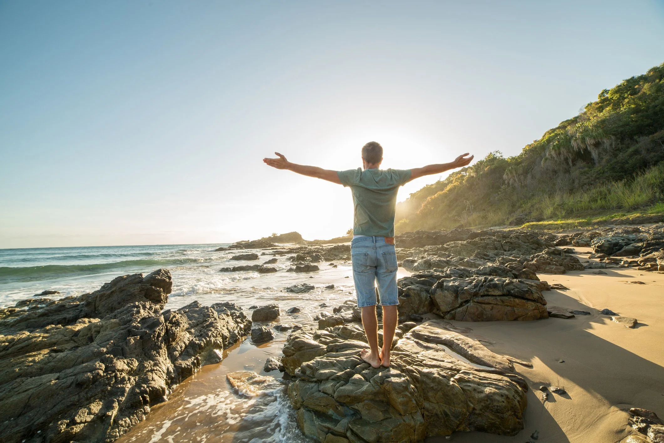 Person standing barefoot on rocks at beach with arms outstretched towards the sun, ocean waves, sandy shore, and green hillside in background.