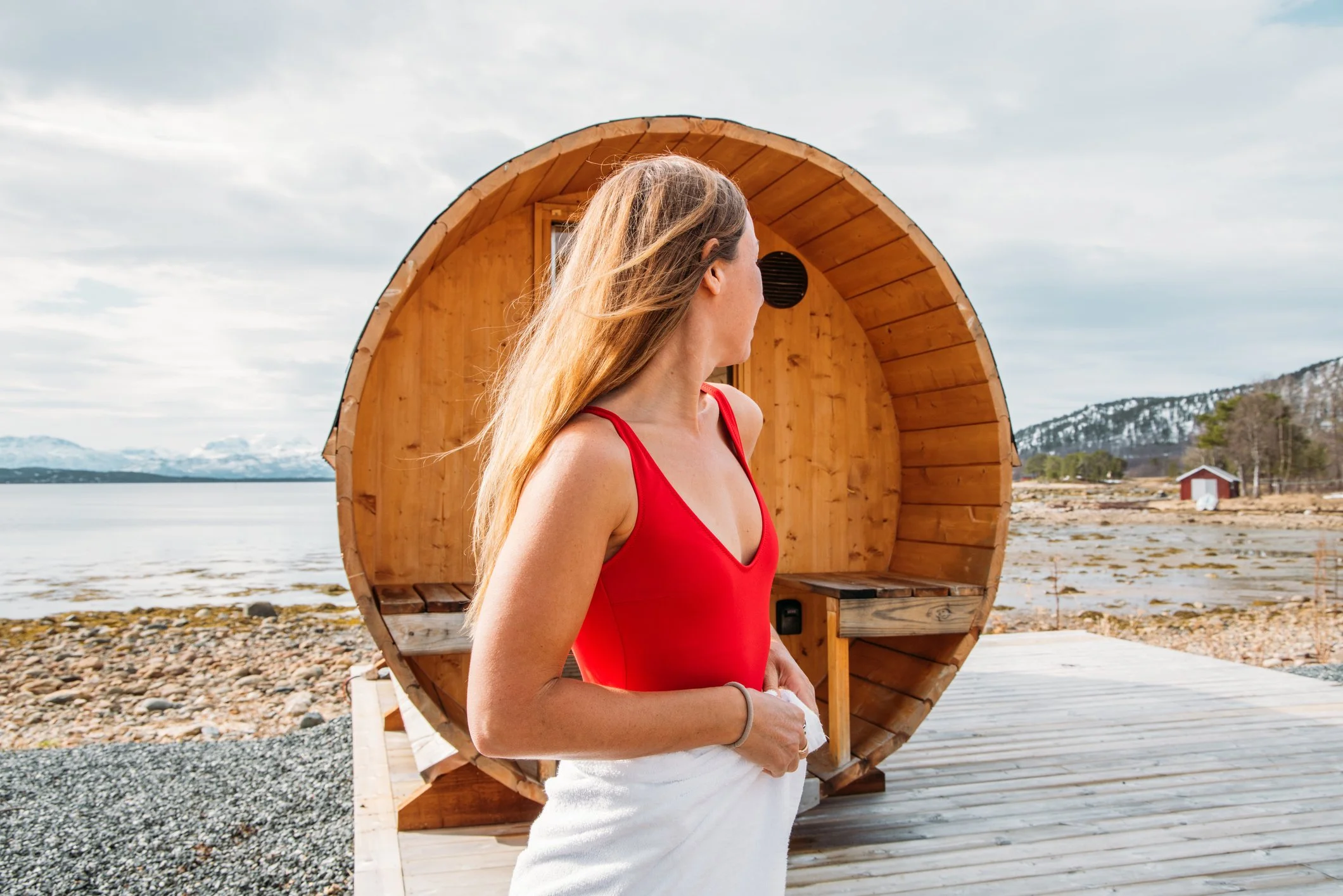 Woman in a red swimsuit with a towel over her waist, standing outside near a wooden barrel sauna by the water with mountains in the distance.