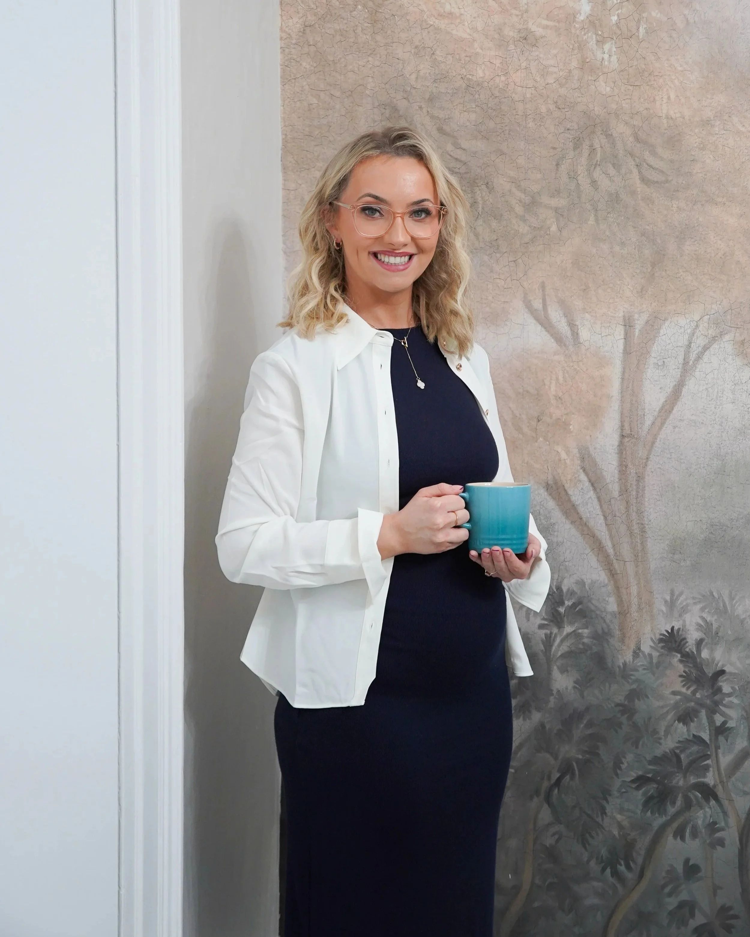 A smiling woman with blonde curly hair and glasses holding a teal mug, wearing a black dress and white blazer, standing in front of a nature-themed mural background.