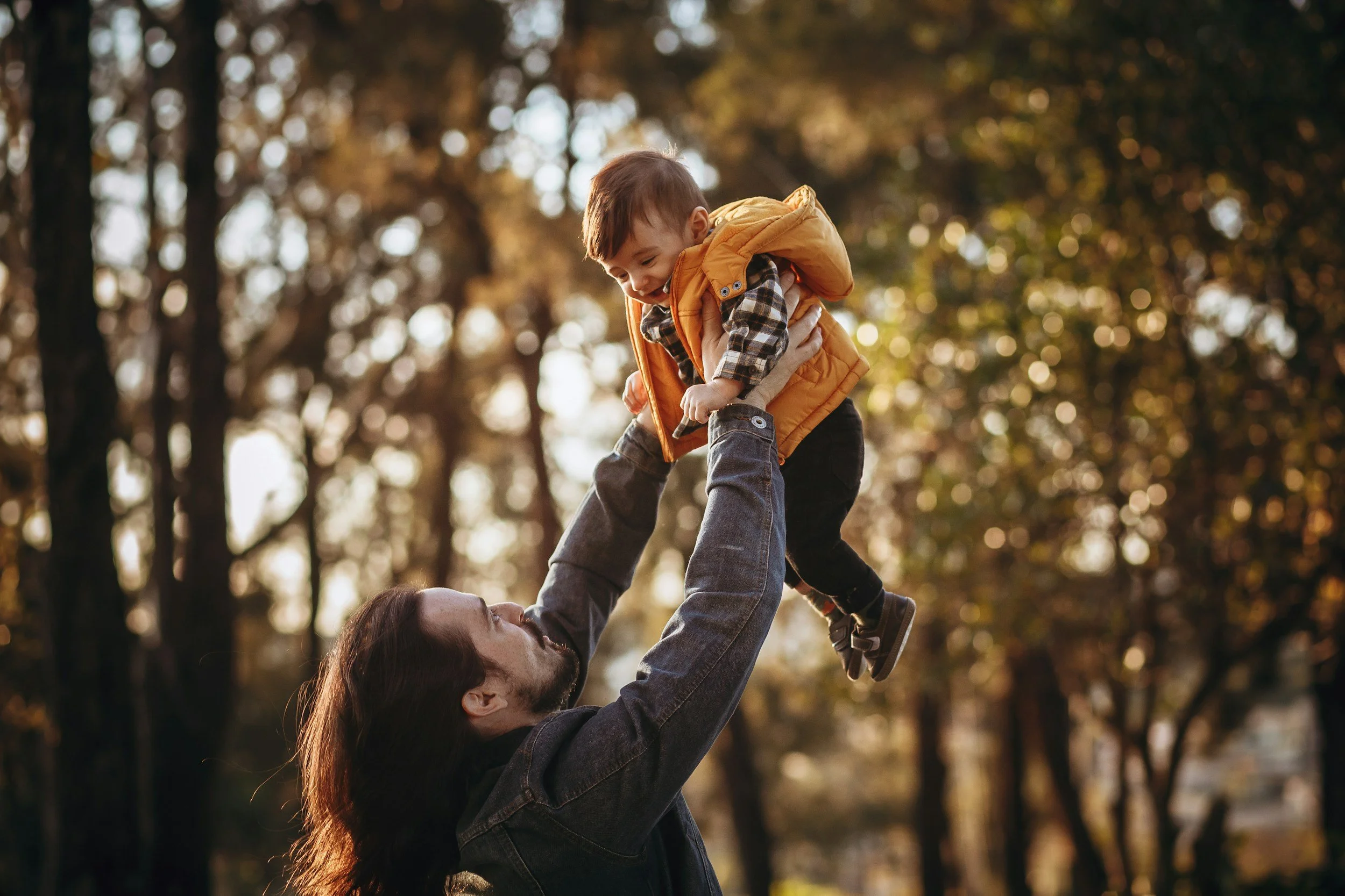A man lifting a young boy in an outdoor park during fall, trees with orange and green leaves in the background.