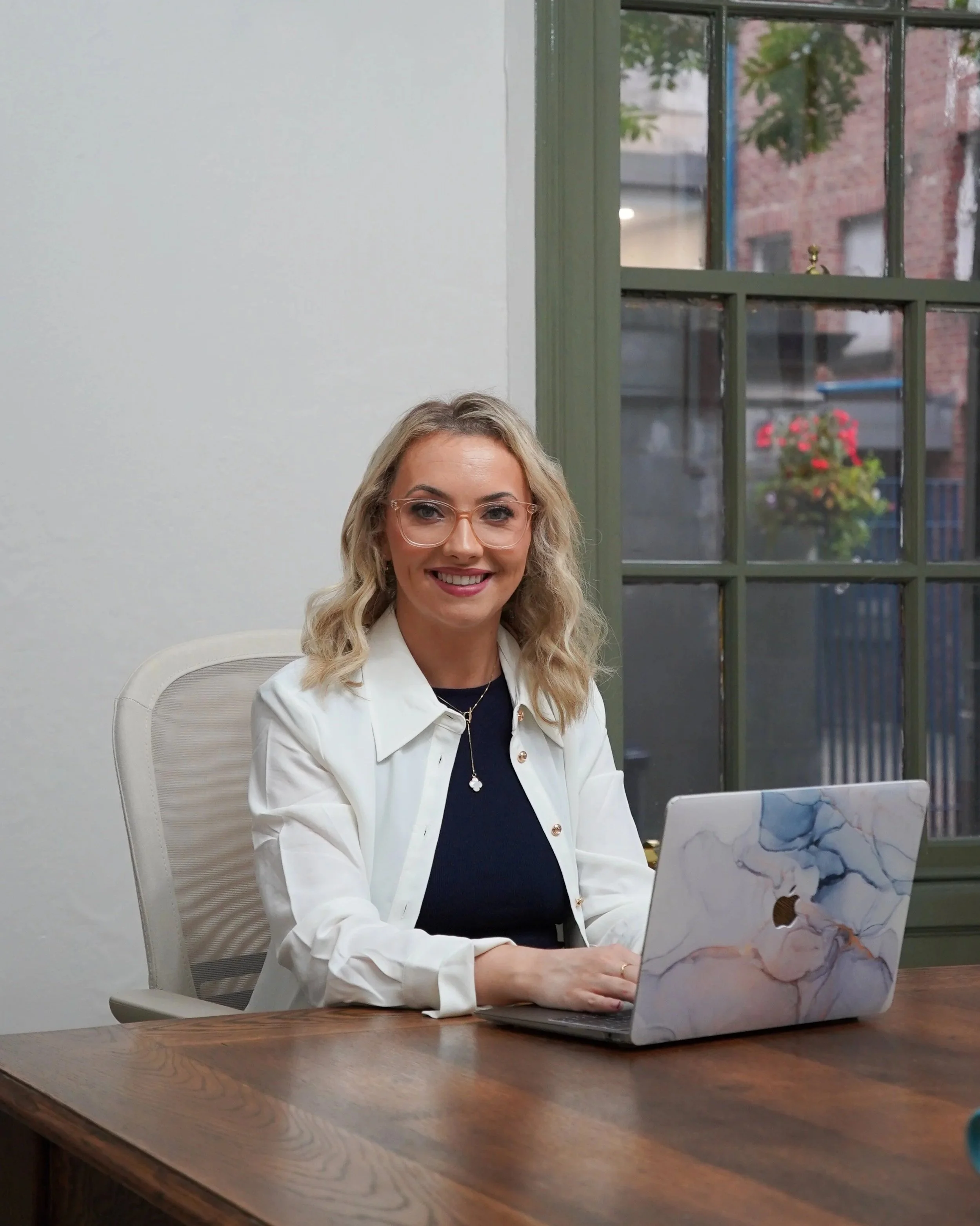 A woman with blonde hair and glasses sitting at a wooden desk with a marble-patterned laptop, smiling, in front of a window with green trim and a view of a brick building and hanging flower basket outside.