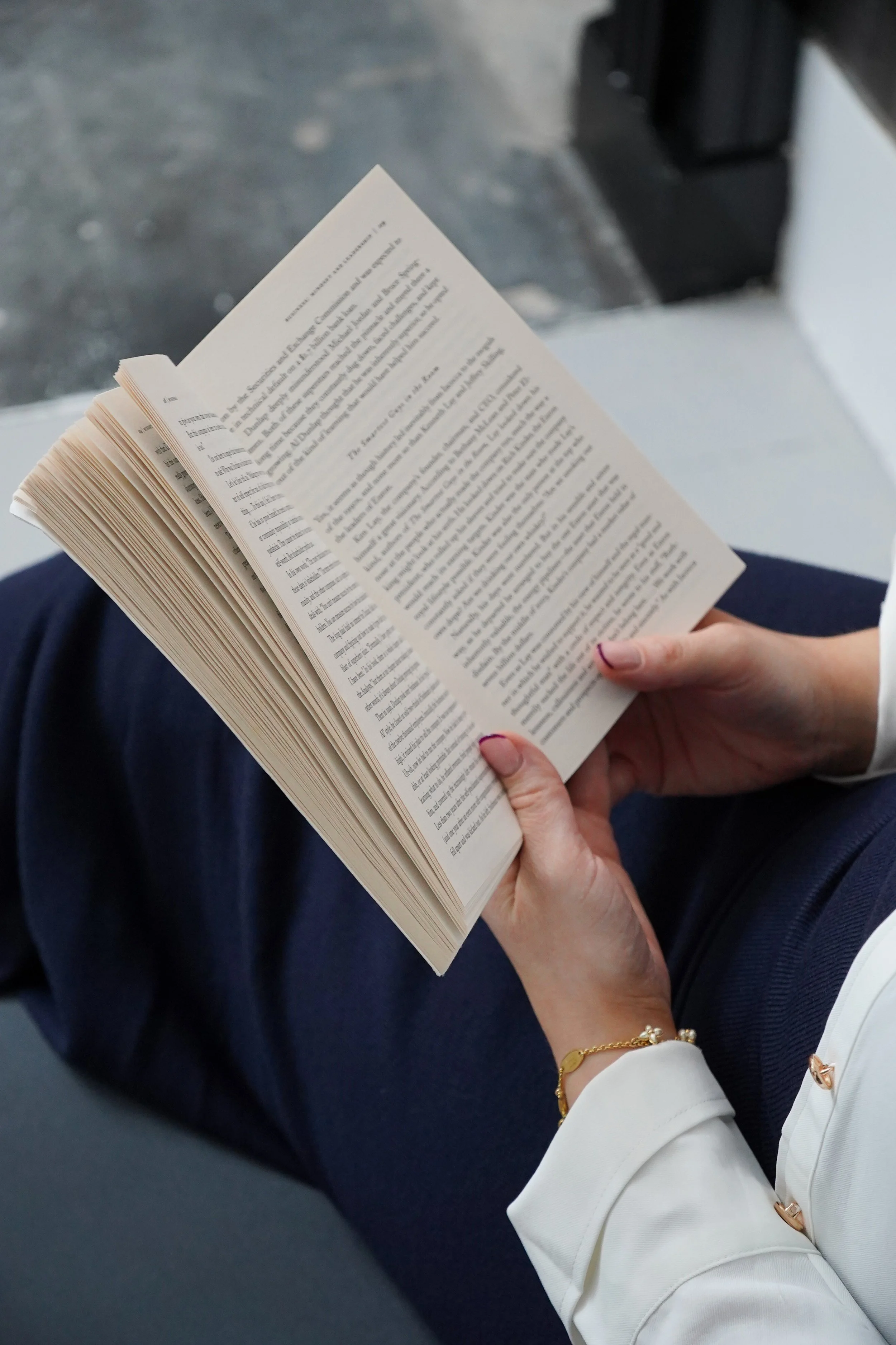 Person holding an open book while sitting down, with a blurry outdoor background.