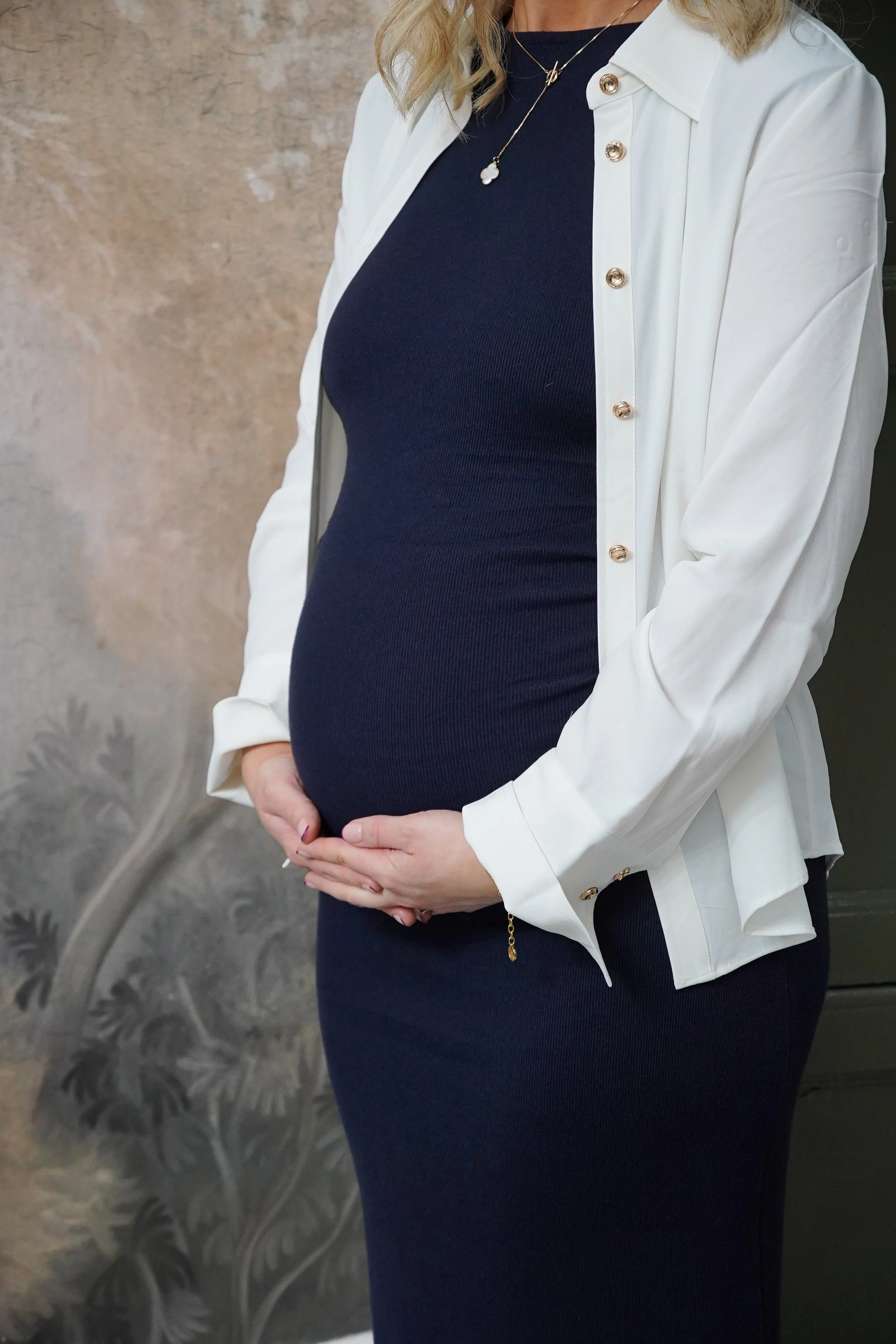 A pregnant woman wearing a navy blue dress and a white button-up shirt, holding her belly.