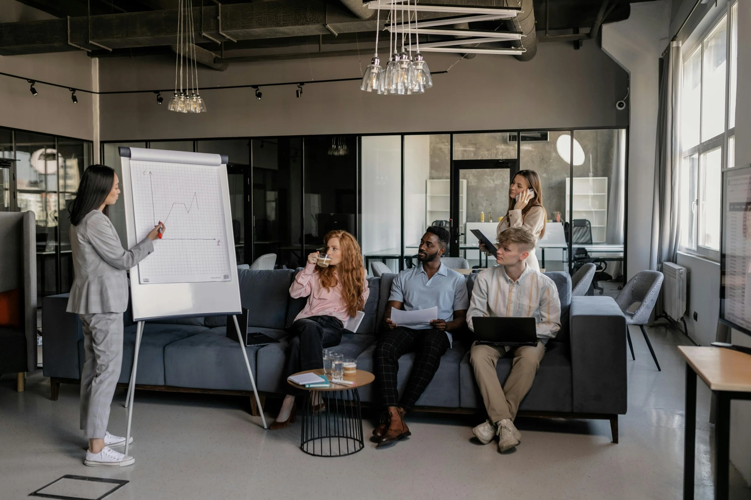 Business meeting in a modern office with five people, one woman presenting data on a flip chart with a line graph, others listening and taking notes, some with laptops and coffee.