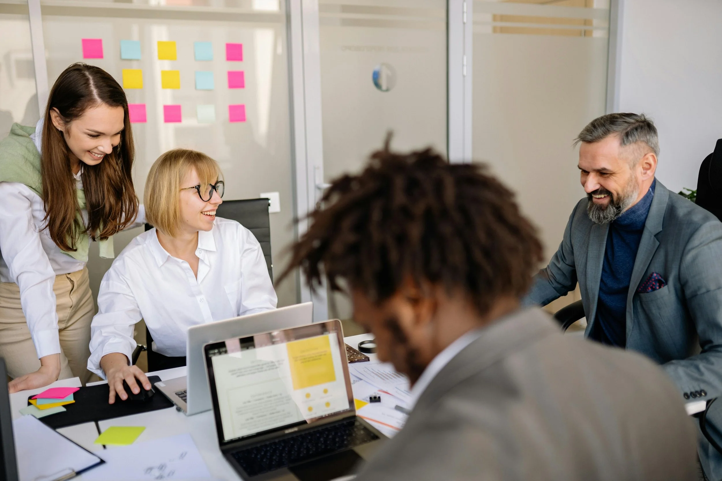 Five people in a business meeting, sitting and standing around a table with laptops, papers, and sticky notes, smiling and engaged in conversation in a modern office.