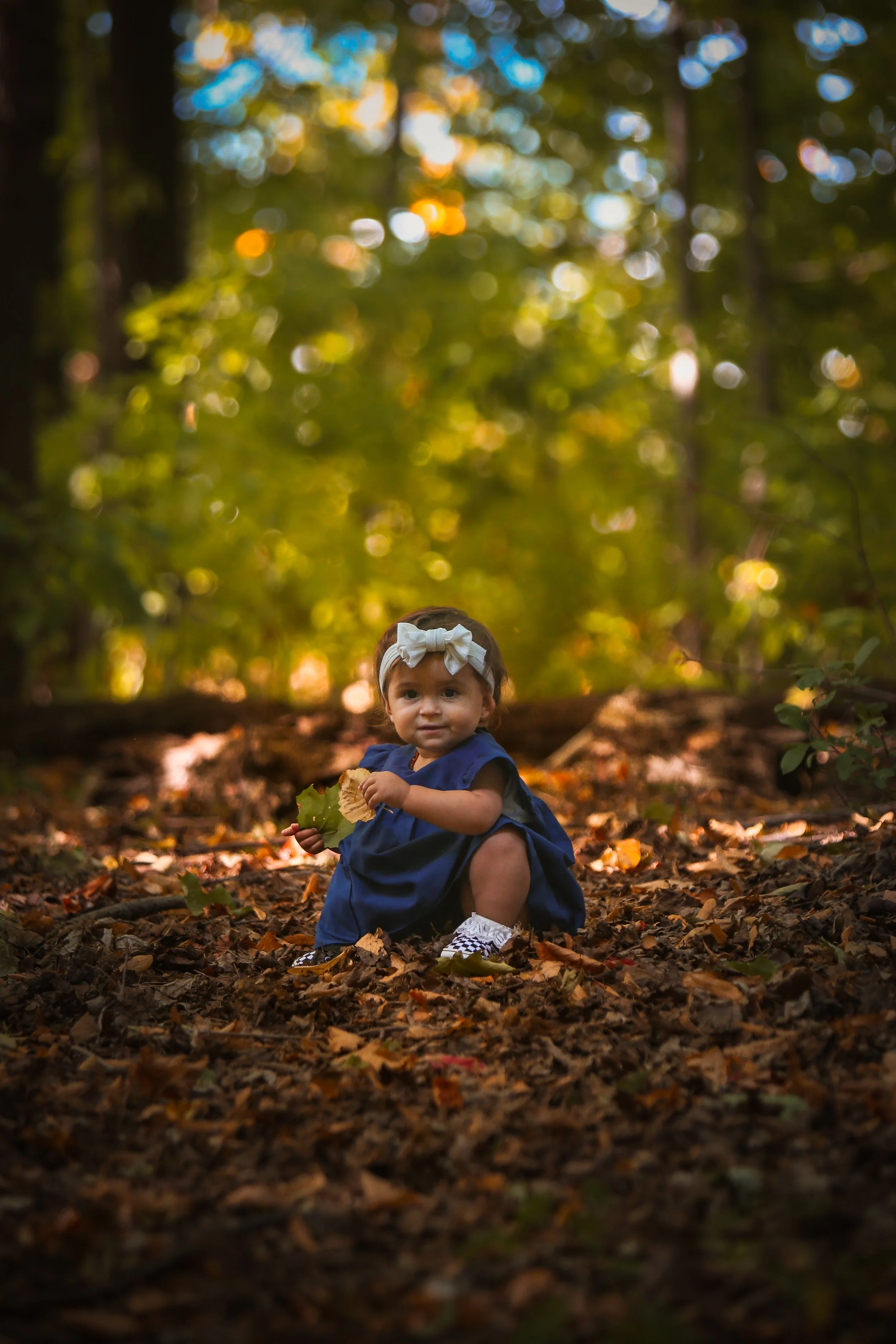 A young girl in a navy dress and white headband squats in a forest, holding leaves, surrounded by fallen autumn leaves, with sunlight filtering through trees in the background.