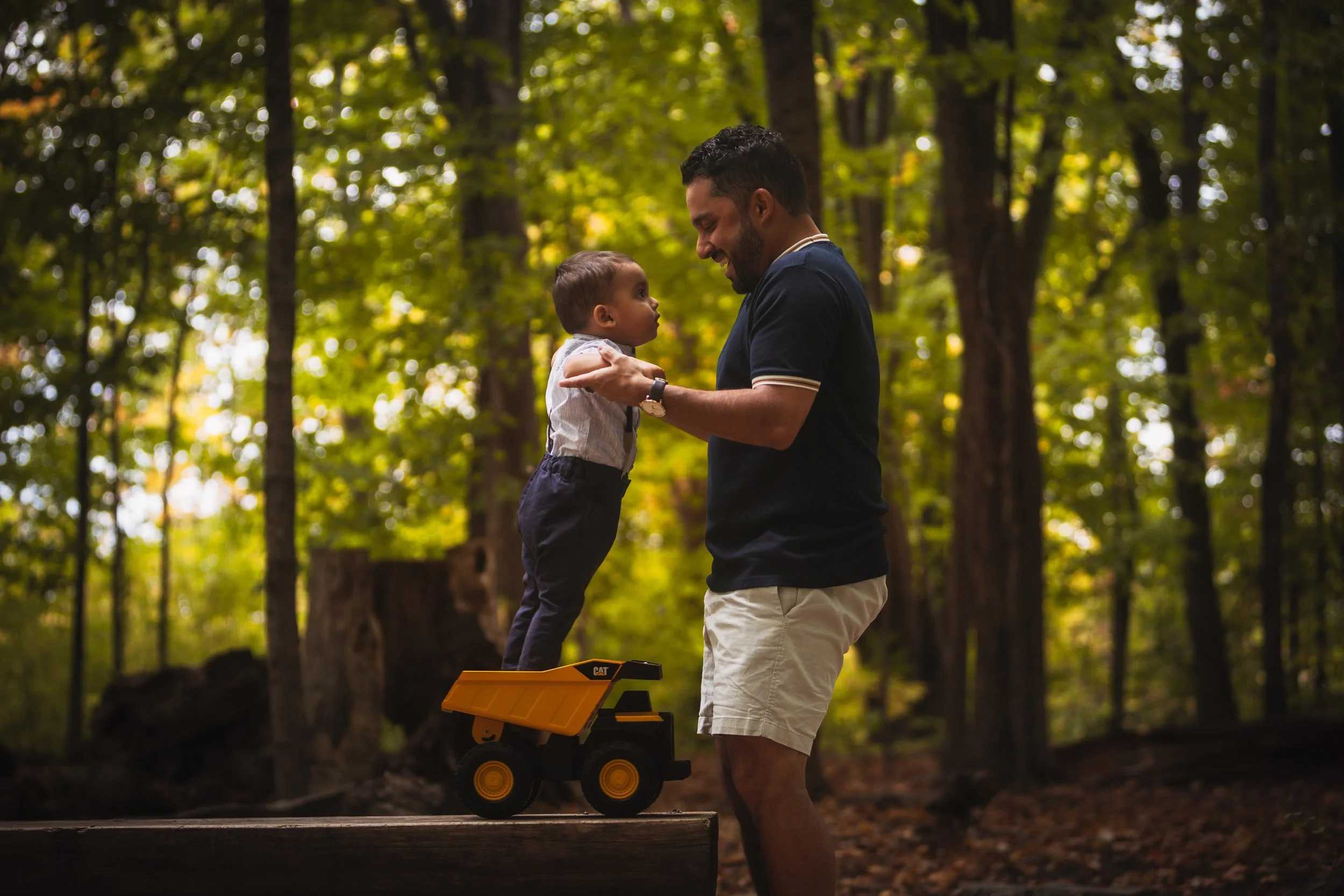 A man and a young boy are in a forest, holding hands, with the boy standing on a toy dump truck. The man is smiling, and the boy is looking at him, both enjoying their time outdoors.