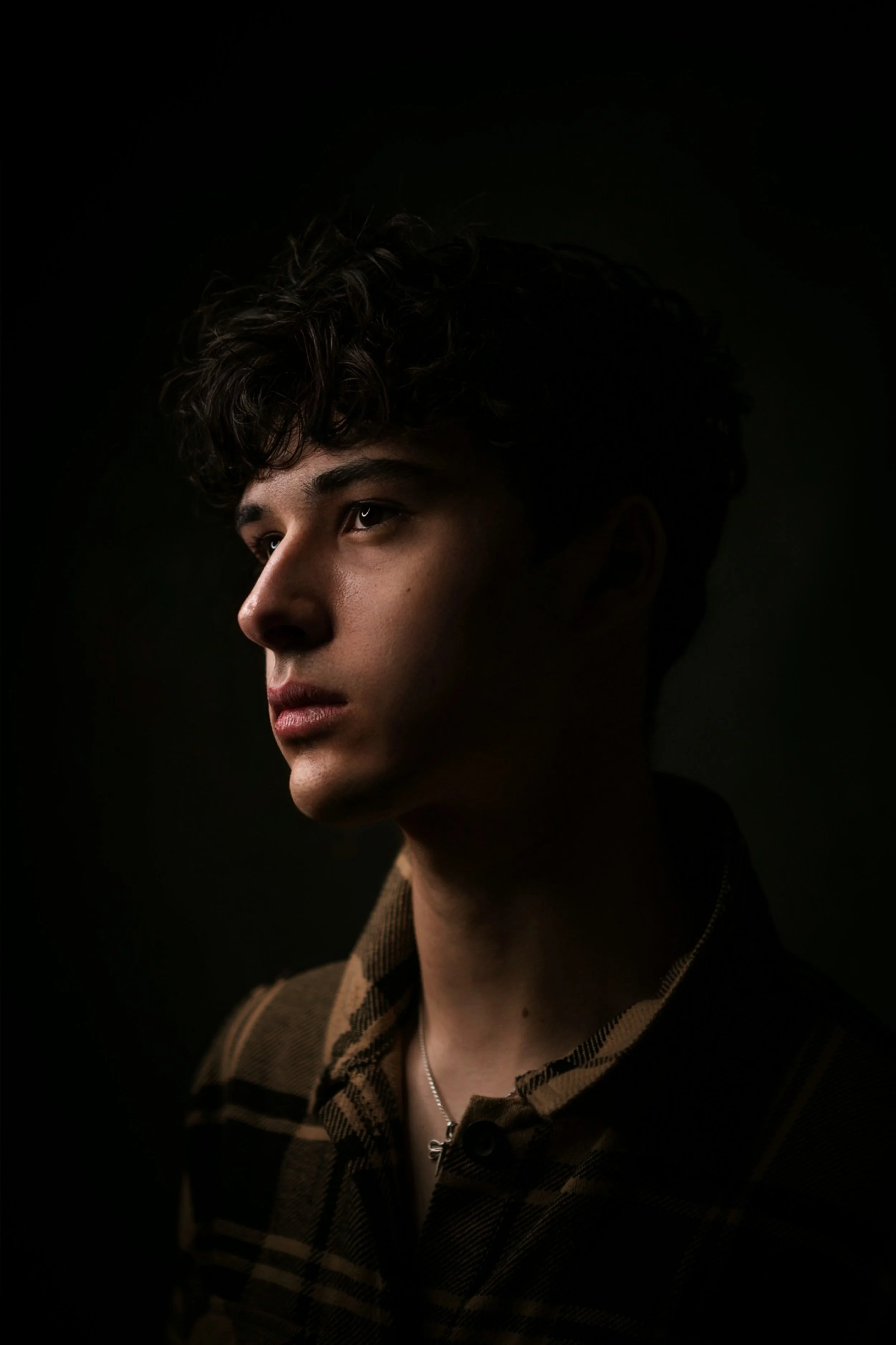 Portrait of a young man with curly hair, wearing a plaid shirt and a silver necklace, against a dark background.
