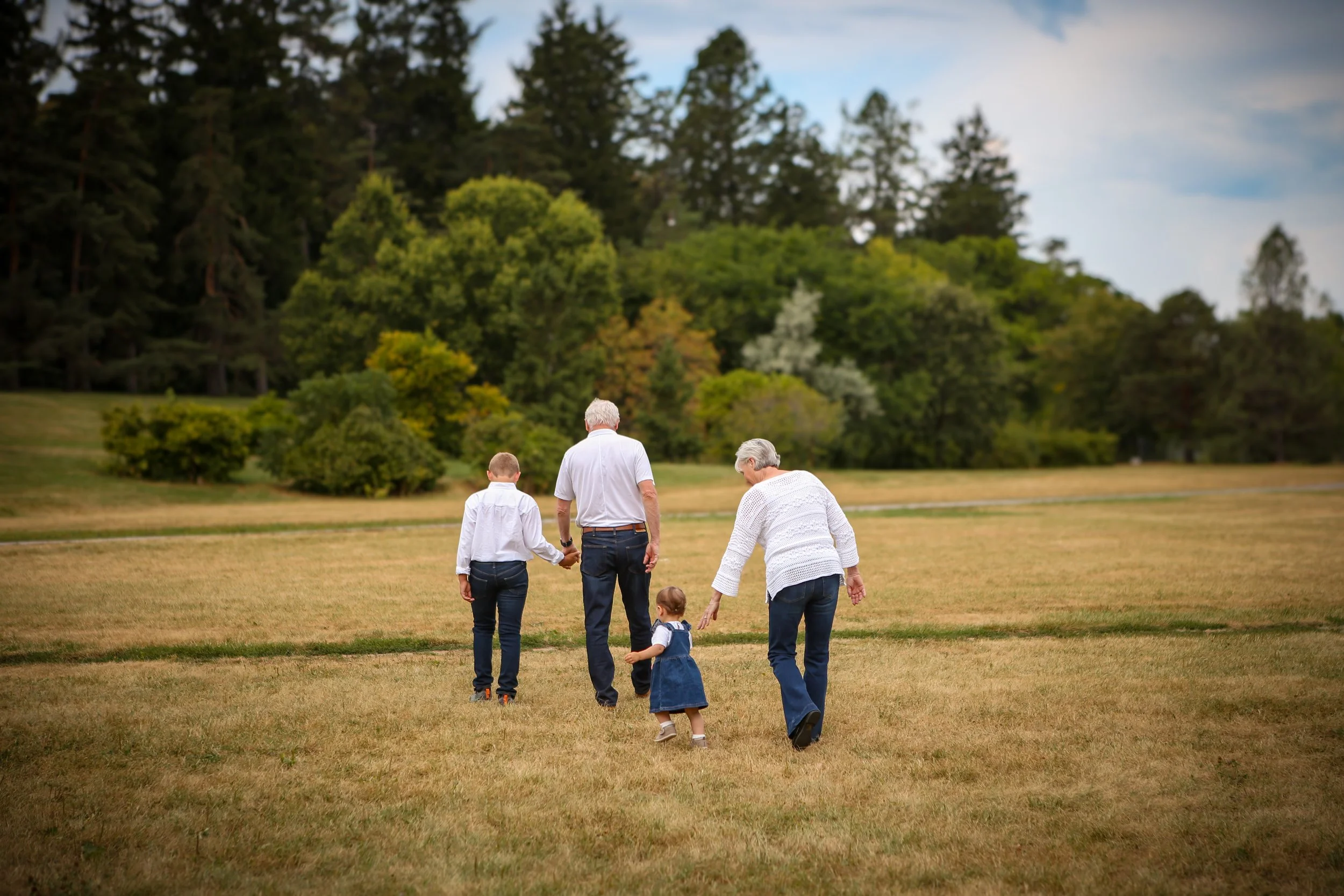 Family of four holding hands and walking on a grassy field with trees and a cloudy sky in the background.