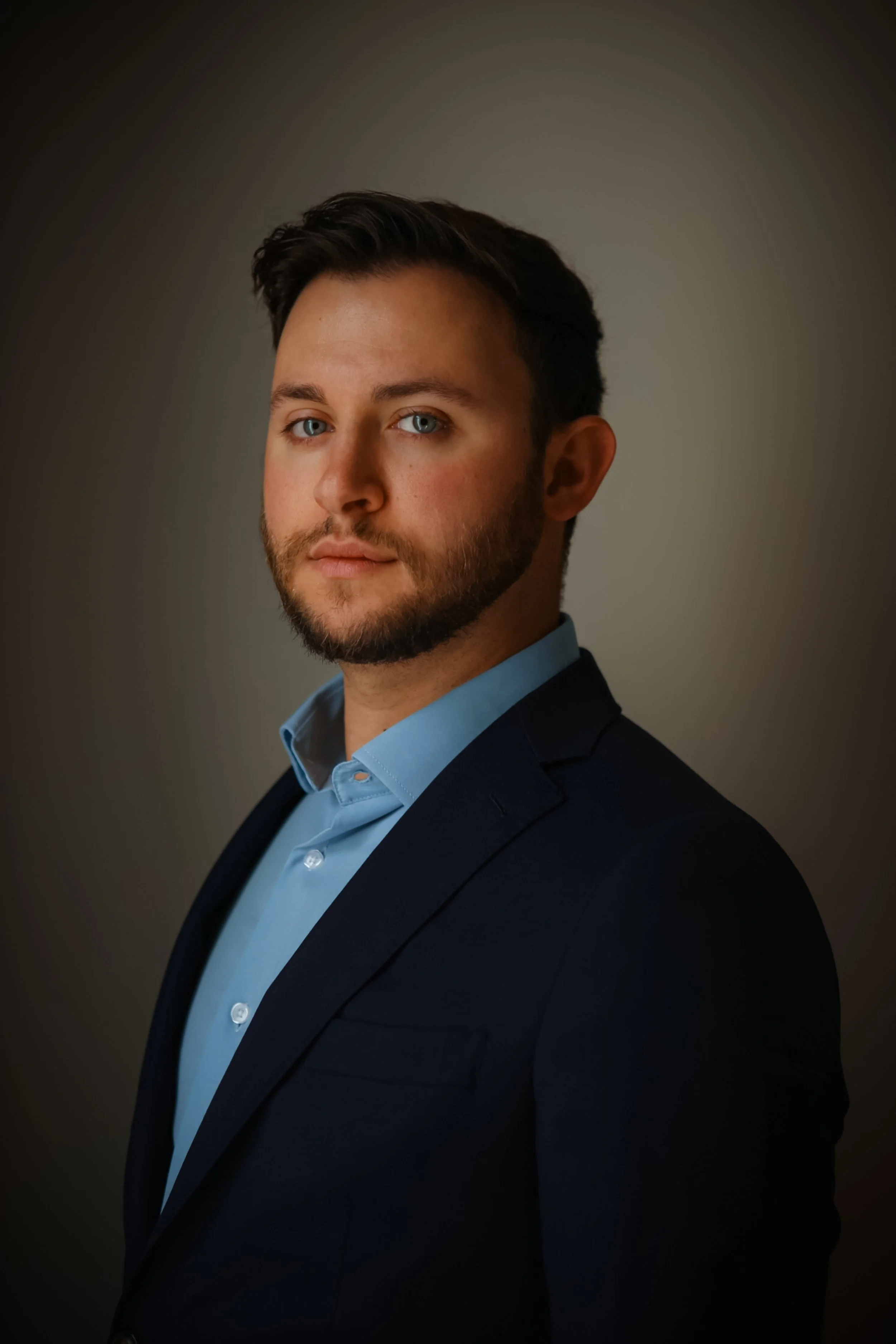 A professional headshot of a young man with short dark hair, blue eyes, and a beard, wearing a navy blazer and light blue dress shirt, against a neutral gray background.