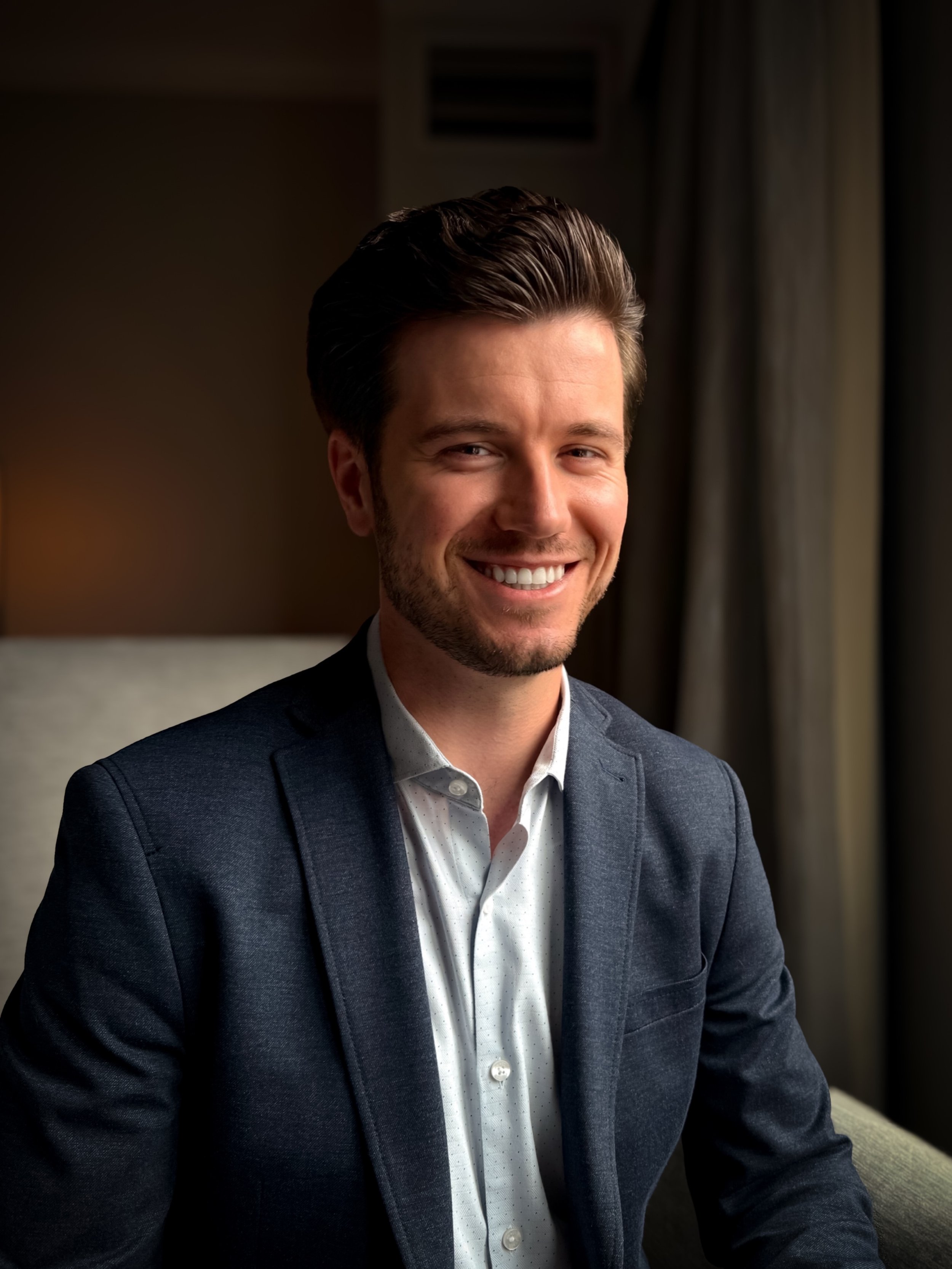 Portrait of a young man with dark hair, wearing a dark blazer and a white button-up shirt, smiling.