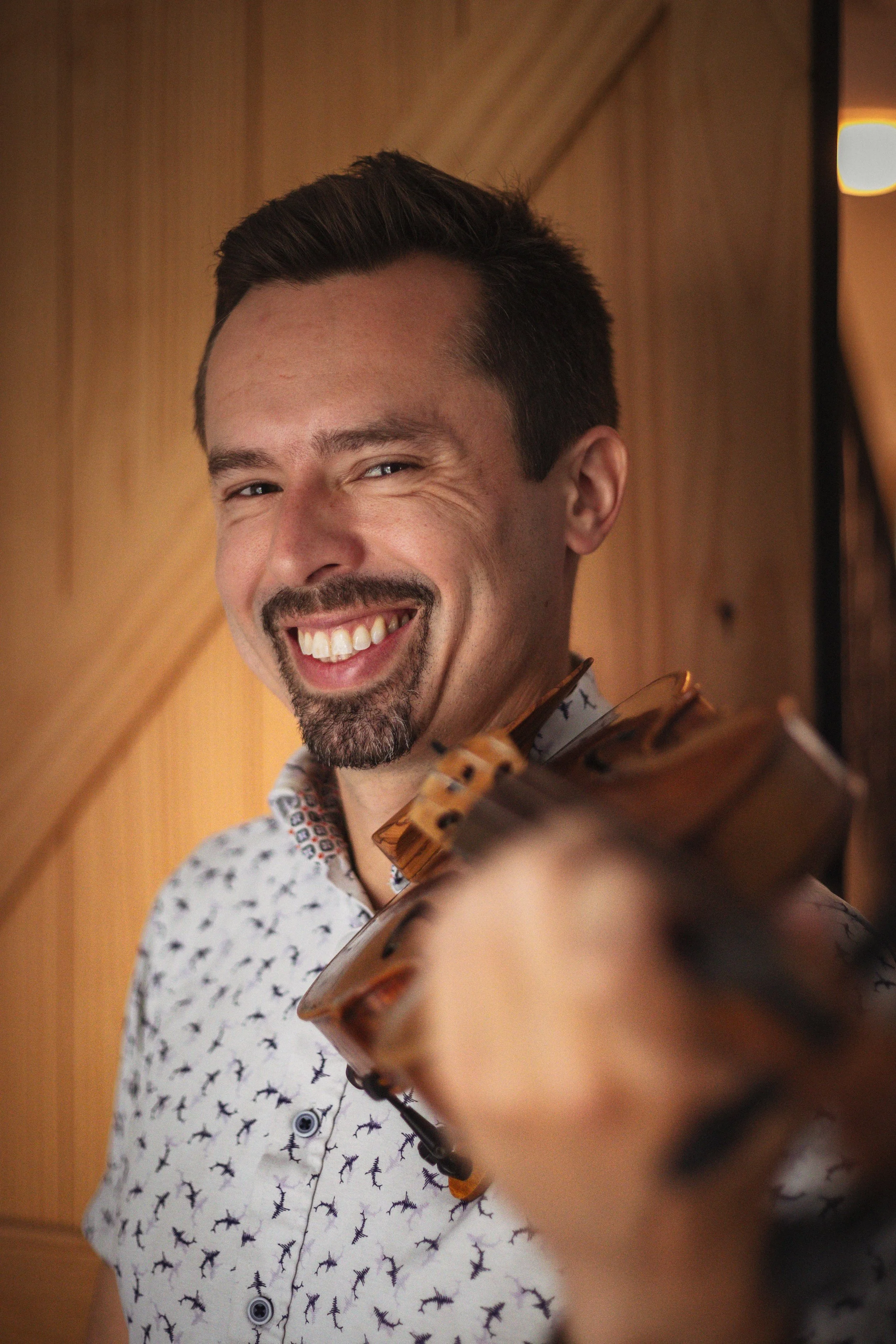 A smiling man with a beard and mustache holding a violin, indoors with wooden walls.