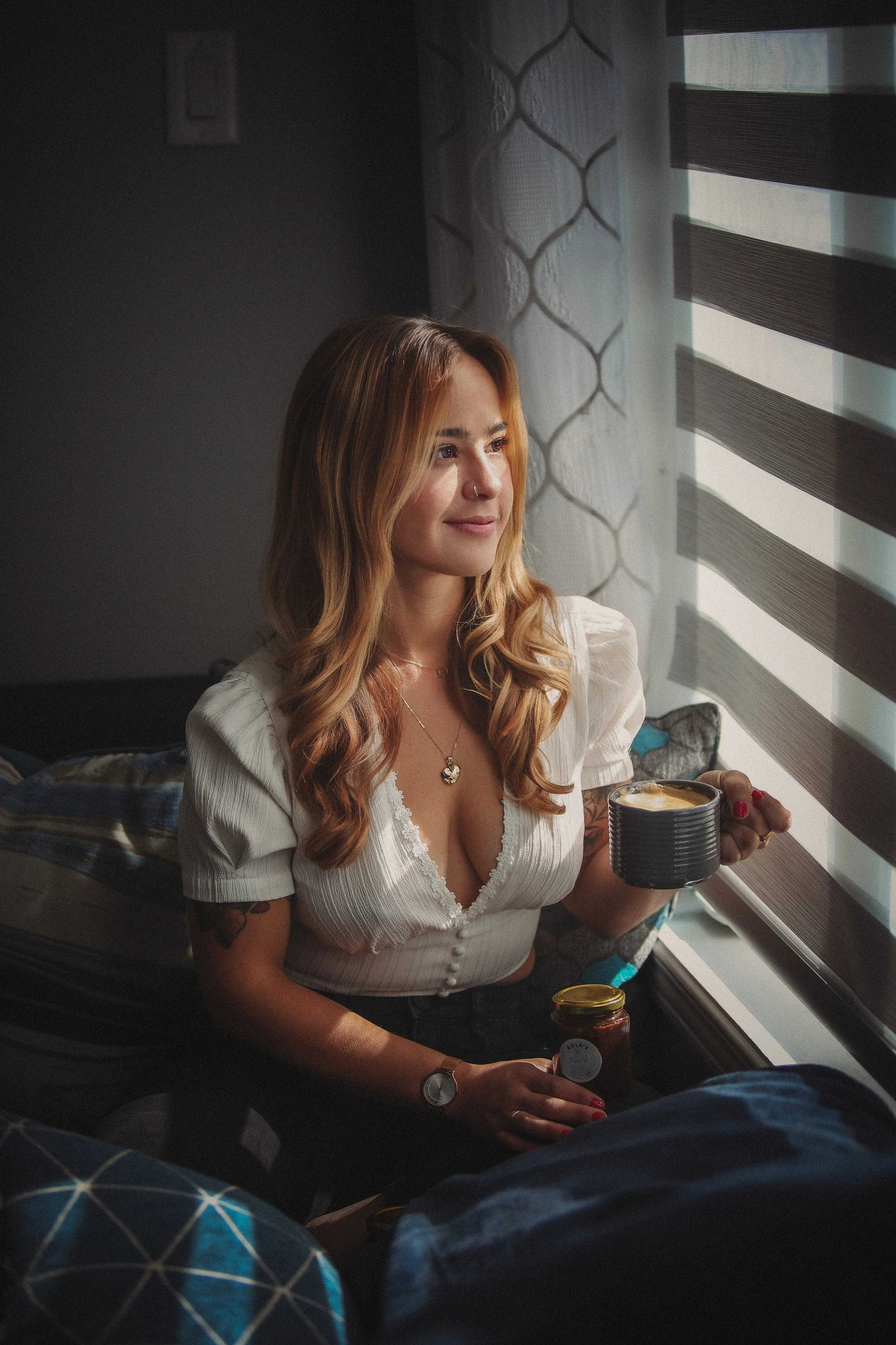 A woman with wavy, reddish-blonde hair sitting by a window with horizontal blinds, holding a mug and a jar of jam, looking out thoughtfully.