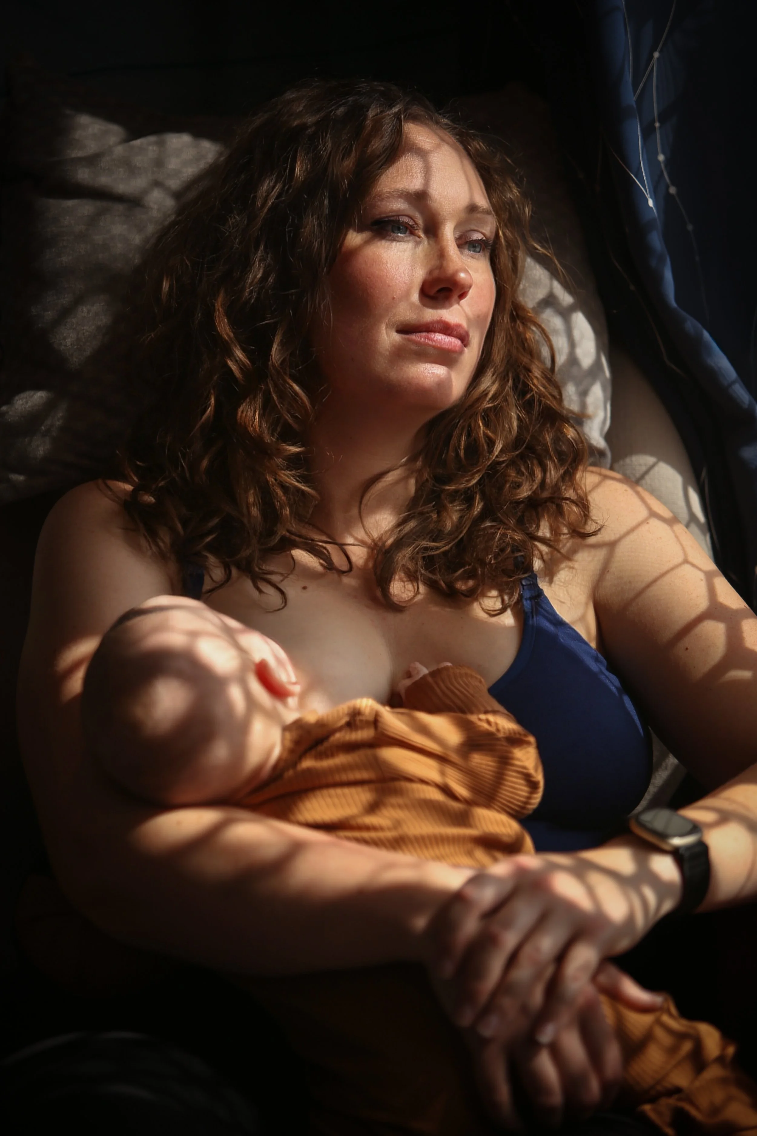 A woman with curly brown hair lying on a bed, her eyes closed, holding a sleeping baby against her chest. Light creates a patterned shadow on her arm.