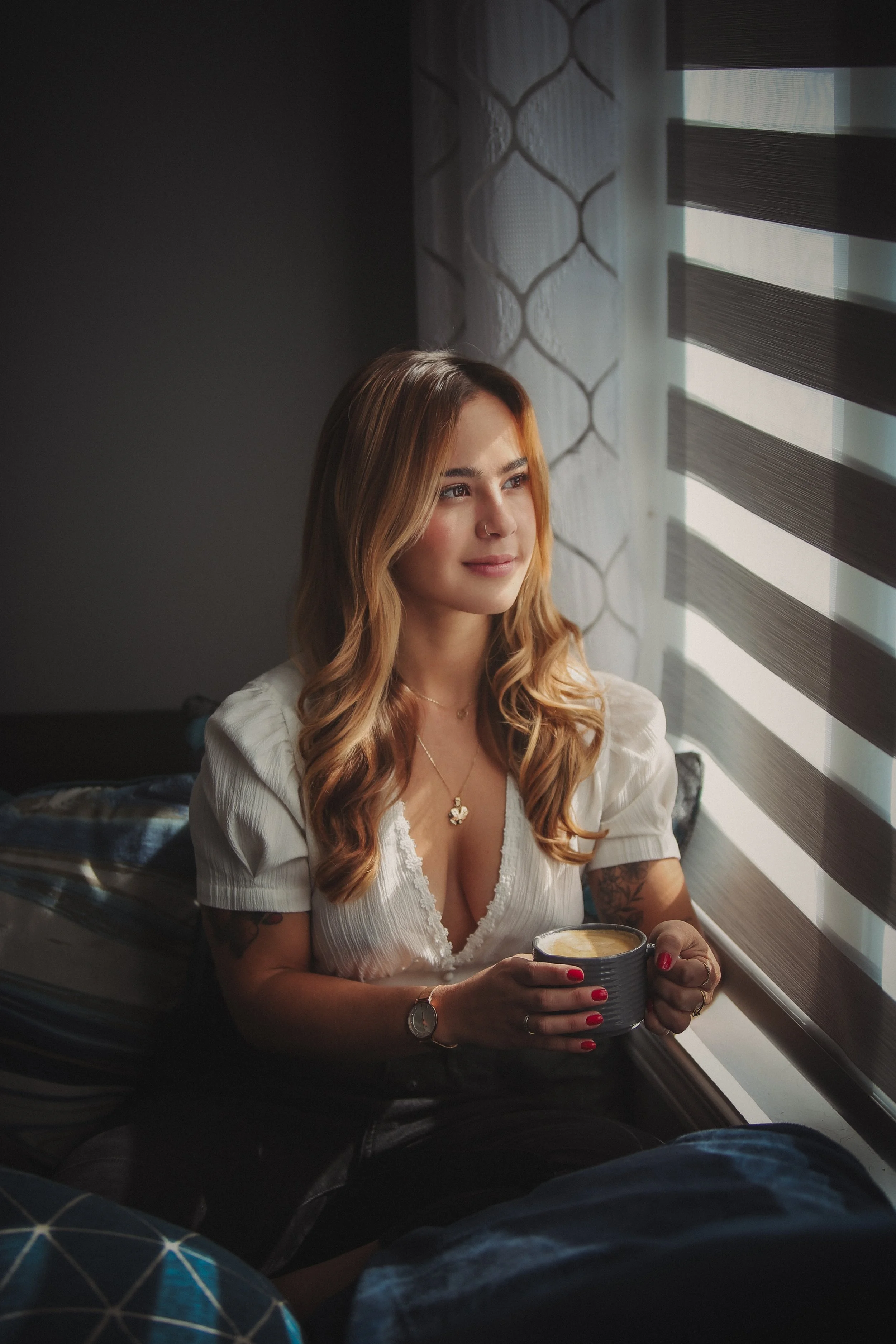 A woman with wavy, light brown hair looks out a window holding a mug, sitting on a bed in a cozy room with patterned curtains and blinds.
