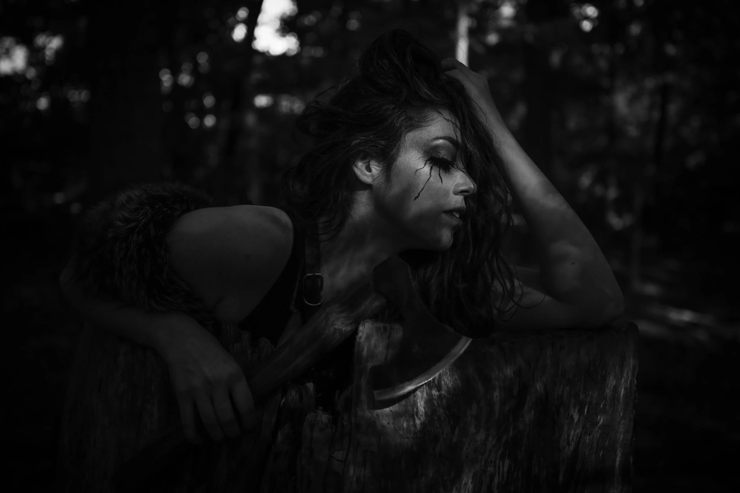 A woman with long, wavy hair leaning on a tree stump in a forest, holding a large axe, with dramatic face makeup resembling a cracked face, in black and white.