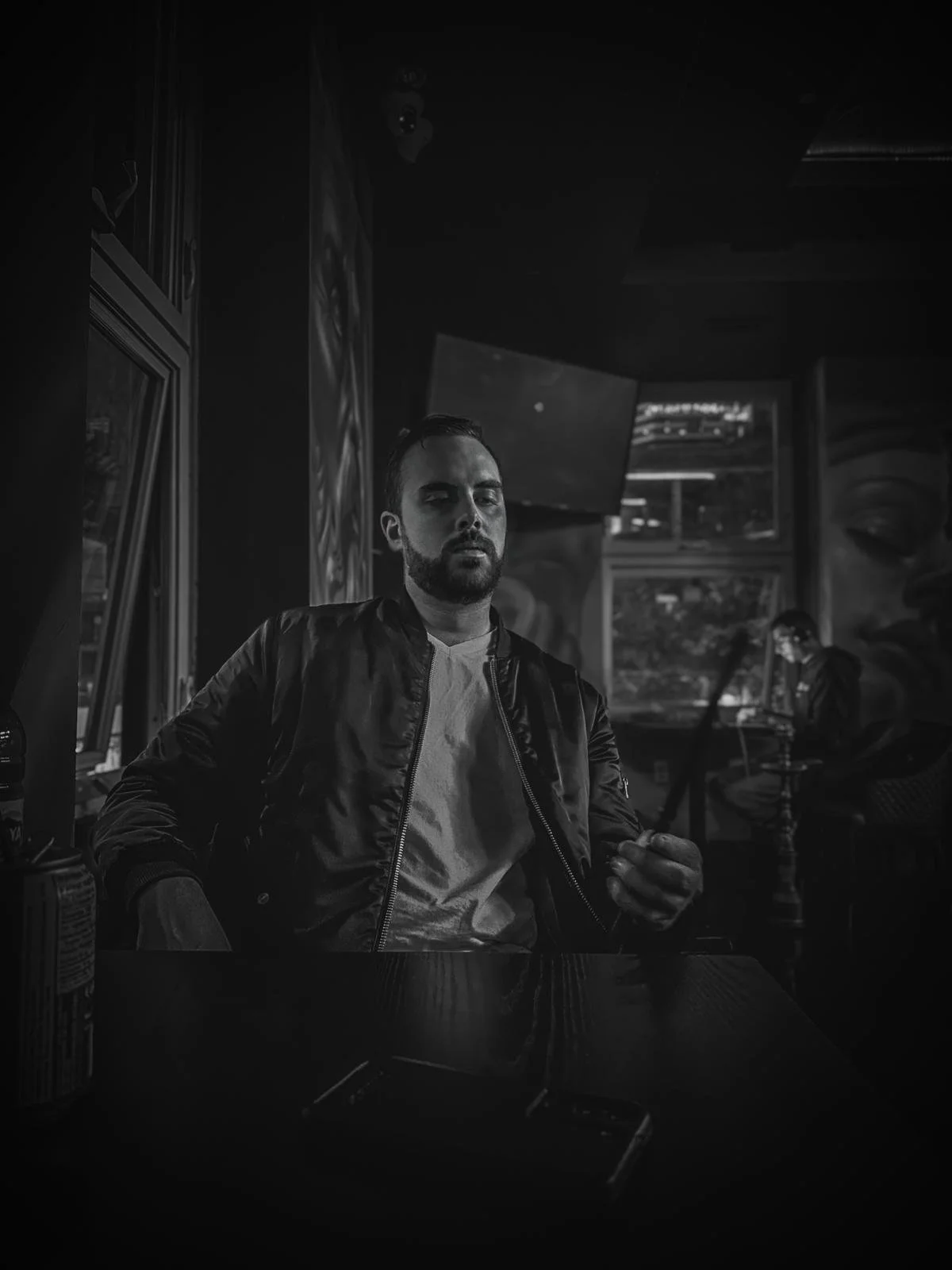 A man with a beard and dark hair sitting at a table in a dimly lit room, wearing a leather jacket and a white shirt, with a magazine and smartphone on the table in front of him.