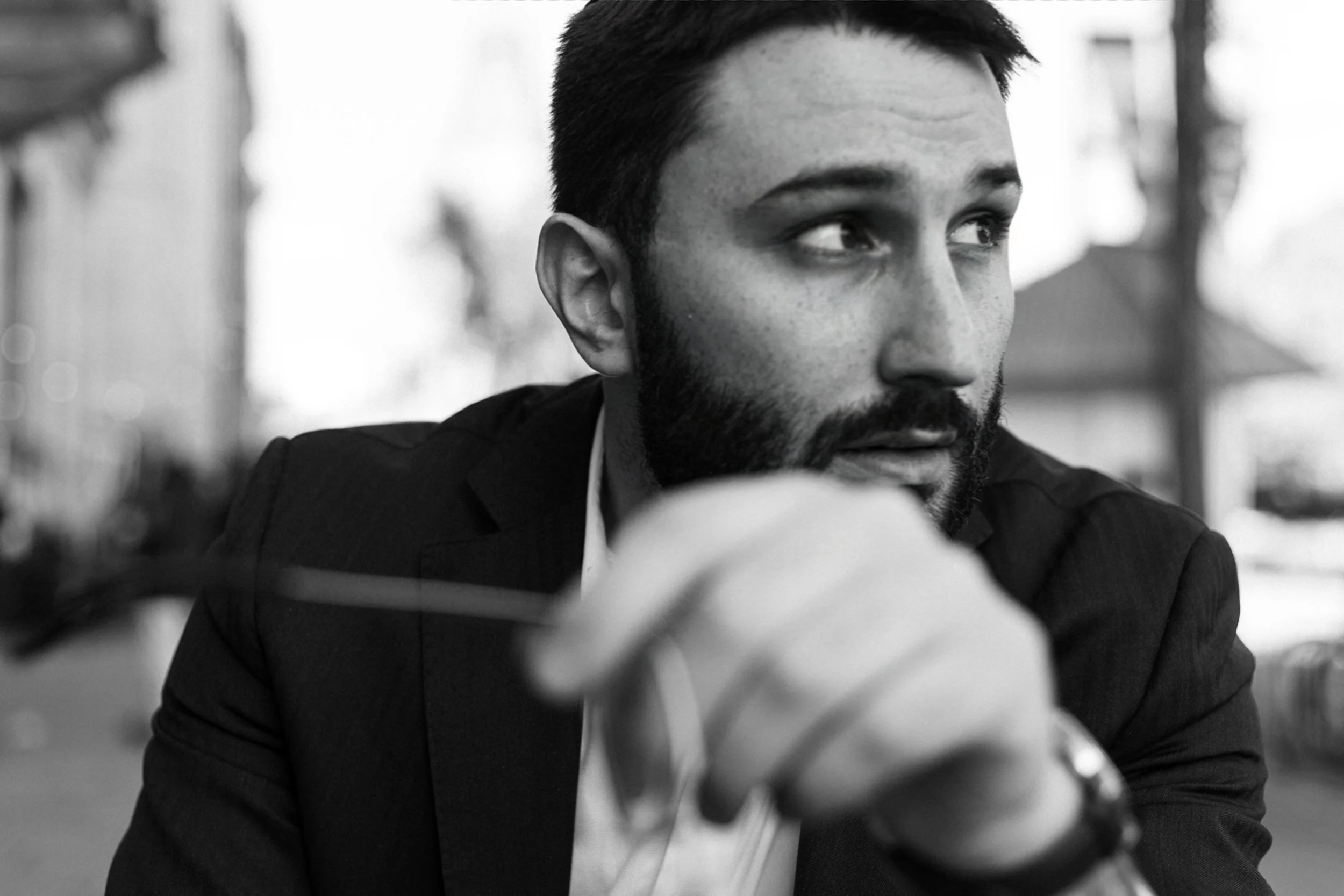 A black-and-white photo of a man with short dark hair and a beard, wearing a suit, looking to the right with a thoughtful expression while holding a pair of eyeglasses in front of him.