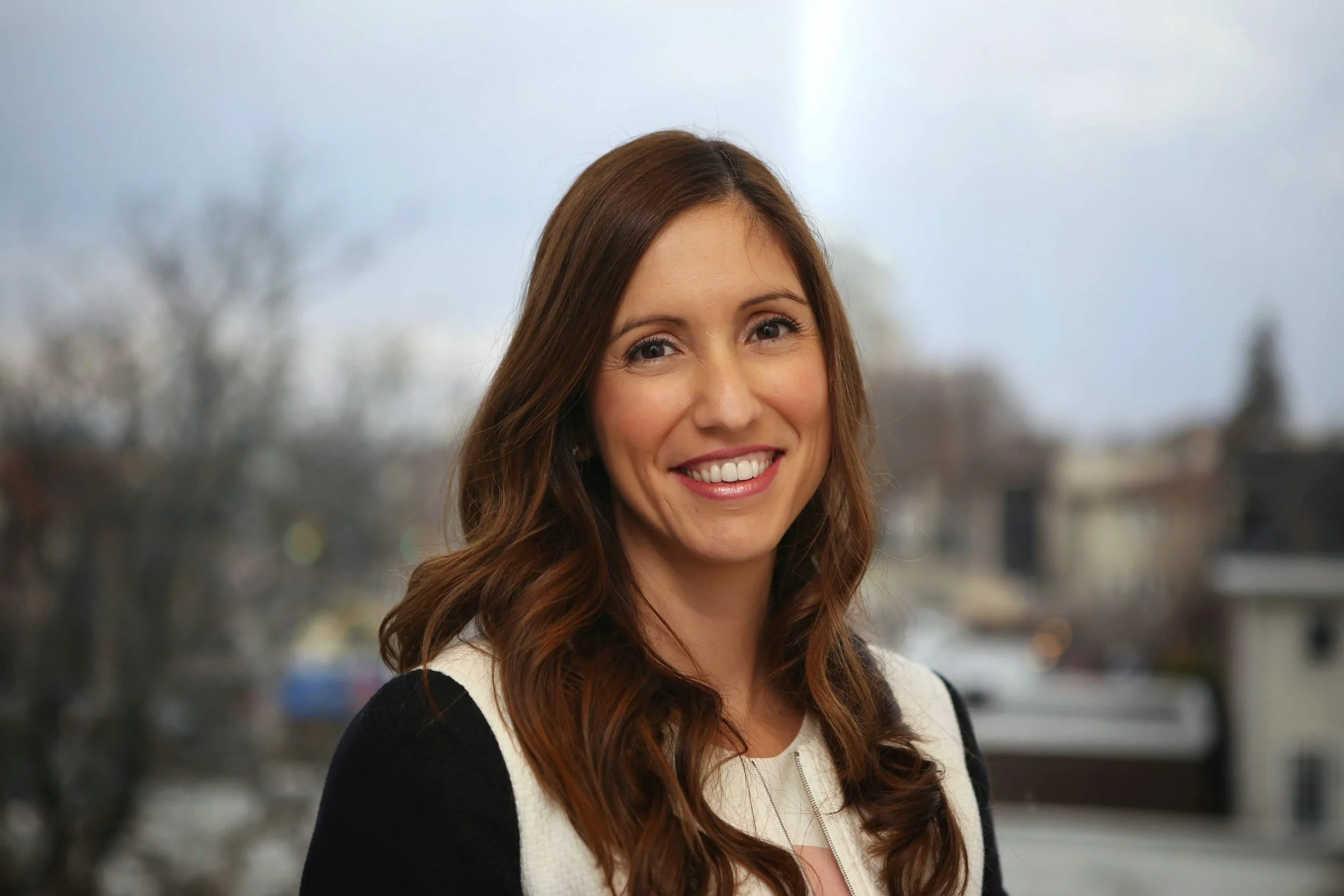 A woman with long brown hair smiling, wearing a black and white outfit, standing outdoors with blurred trees and buildings in the background.