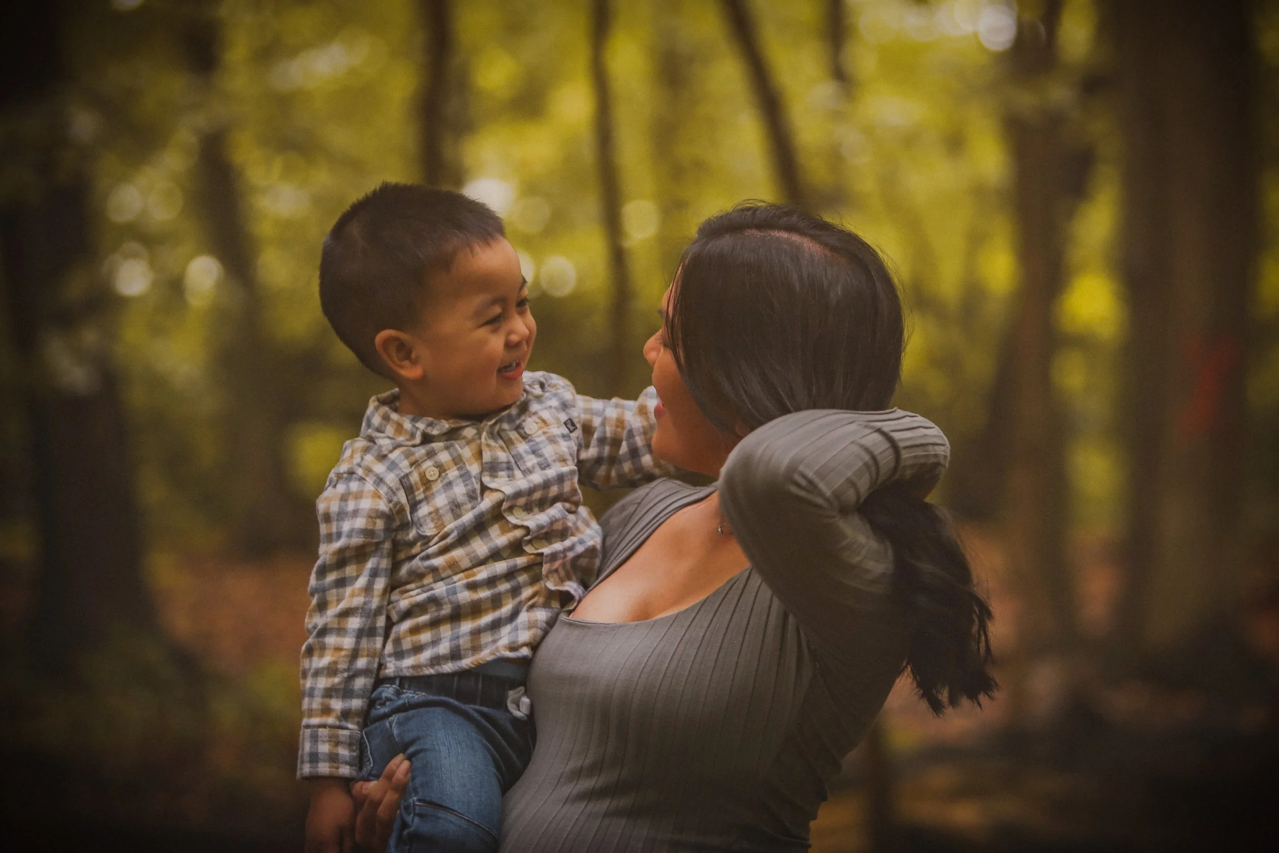A woman lifting a young boy in a forest during autumn, both smiling and looking at each other.