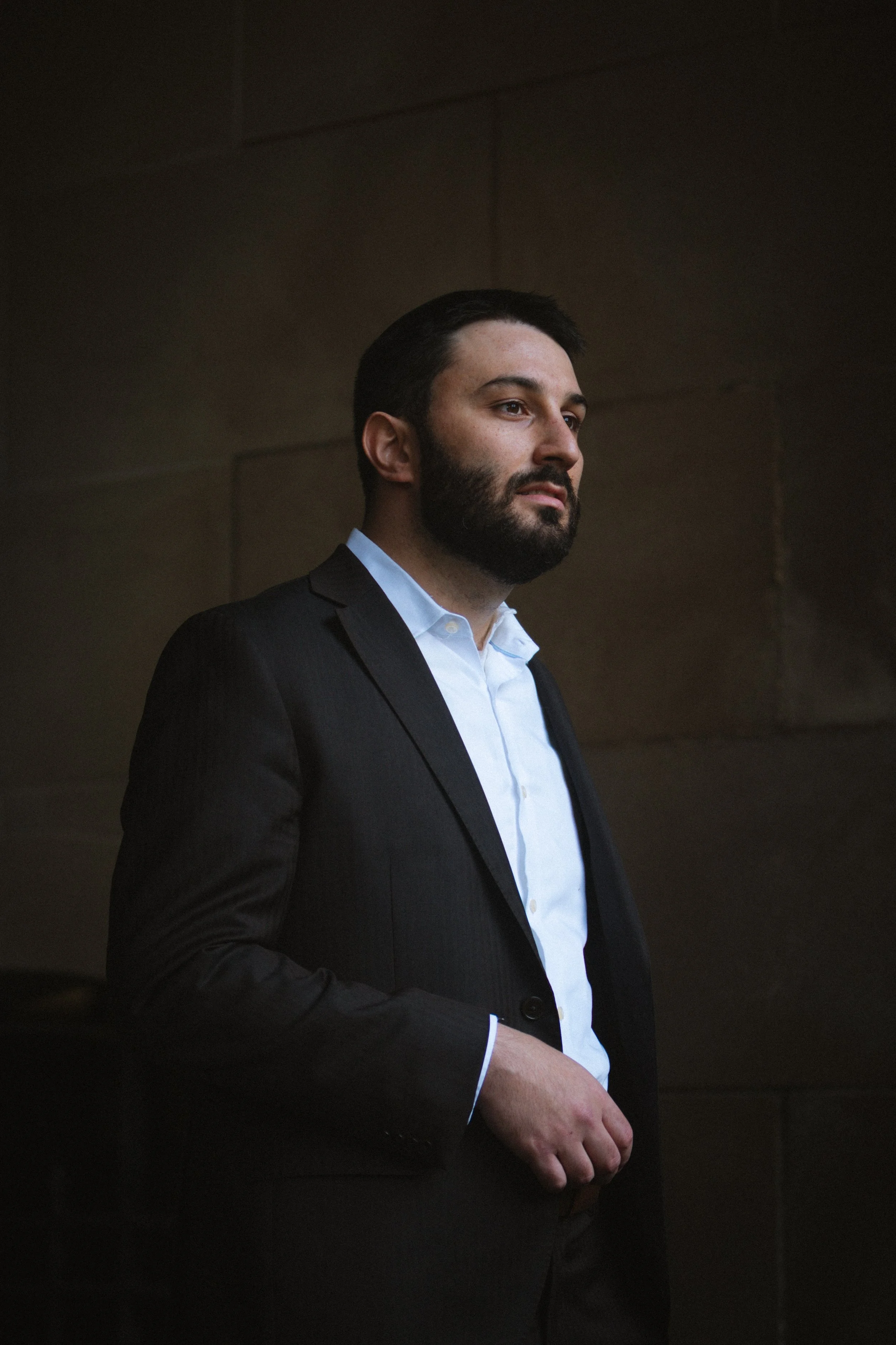 A man with dark hair and a beard wearing a dark suit and light blue shirt, standing with his hand in his pocket against a brown wall.