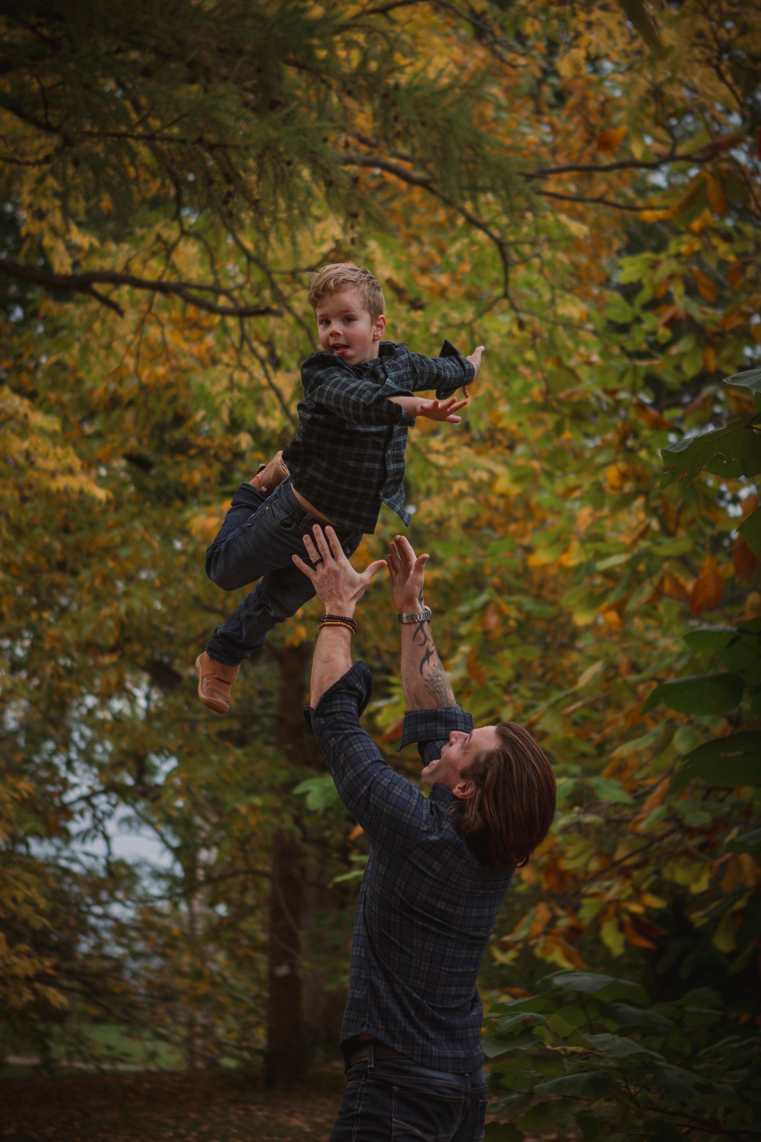 A man lifting a young boy in the air outdoors during fall with autumn leaves in the background.