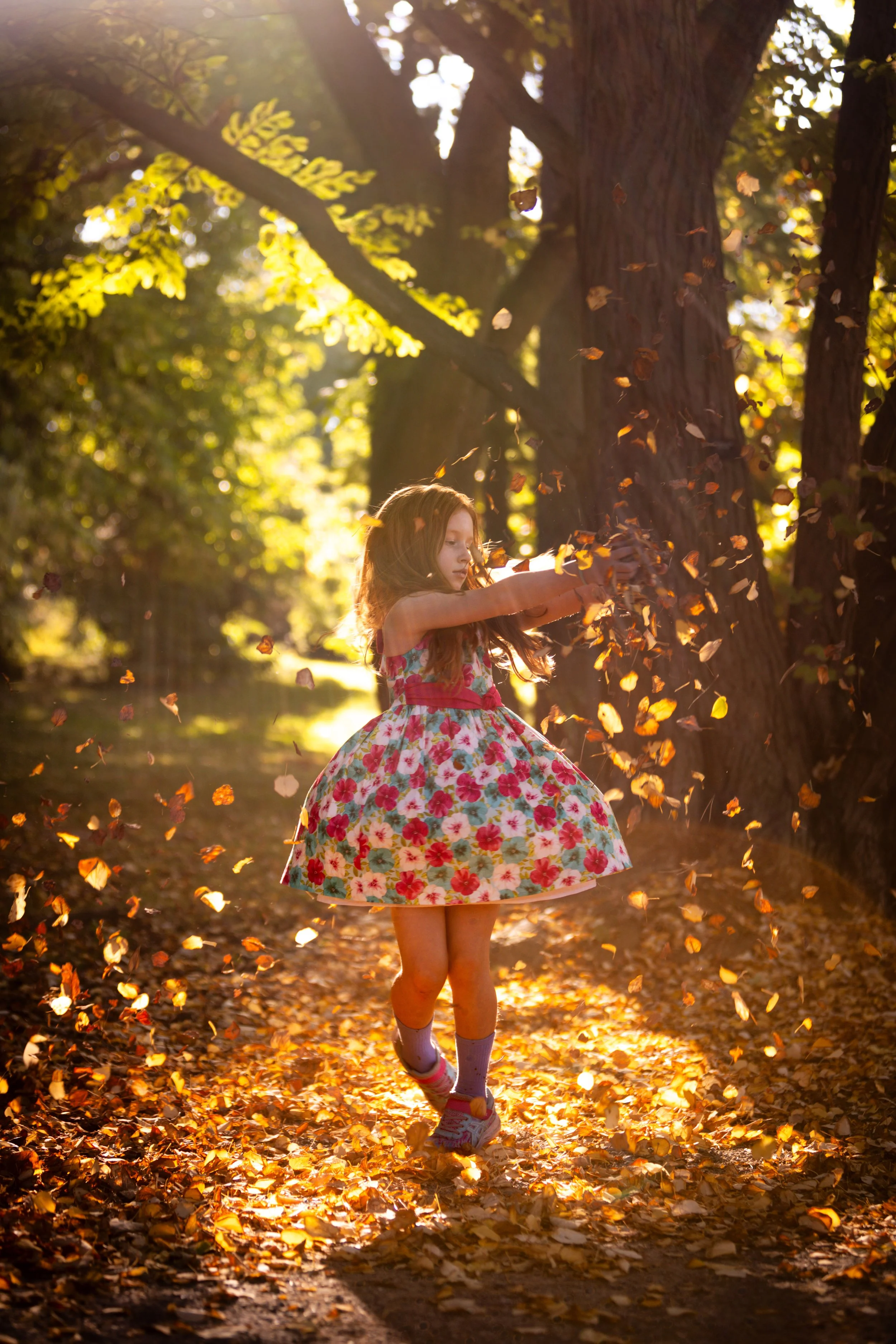 A young girl in a floral dress playing with fallen autumn leaves on a sunlit forest trail.