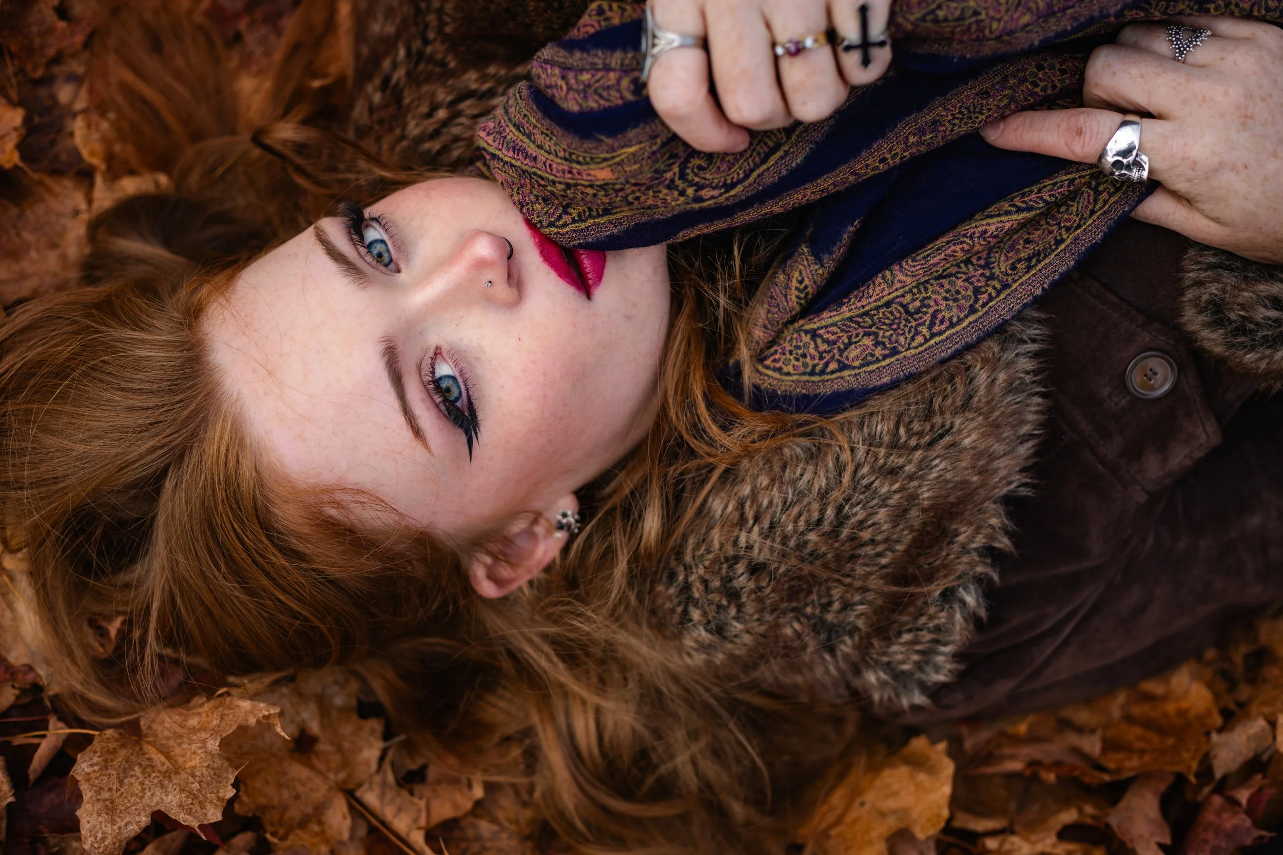 A young woman with red hair and bright blue eyes lying on fallen autumn leaves, wearing a patterned scarf, a brown furry coat, and multiple rings, holding her hand to her face.