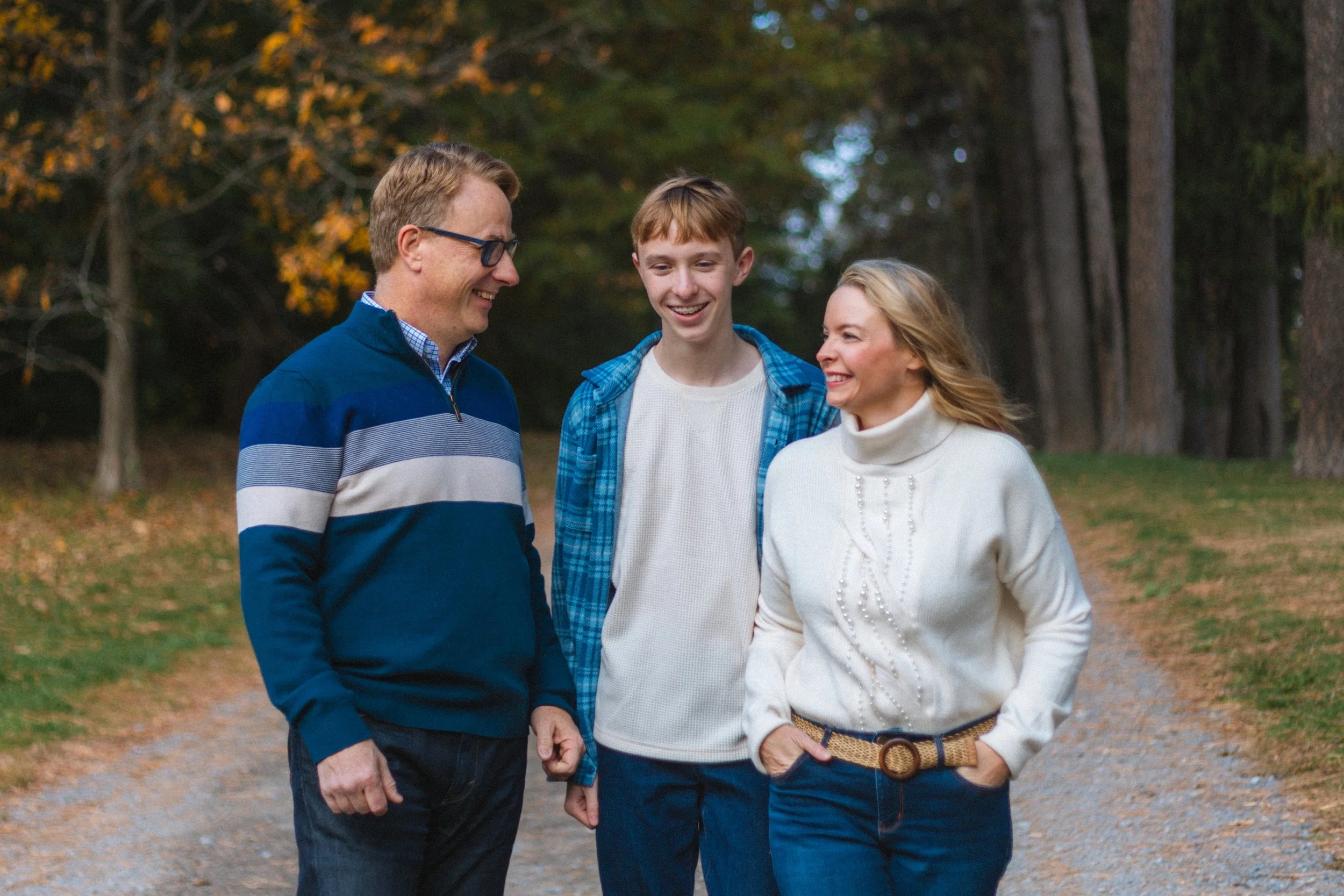 A family of three walking outdoors on a trail surrounded by trees with fall foliage, smiling and talking together.