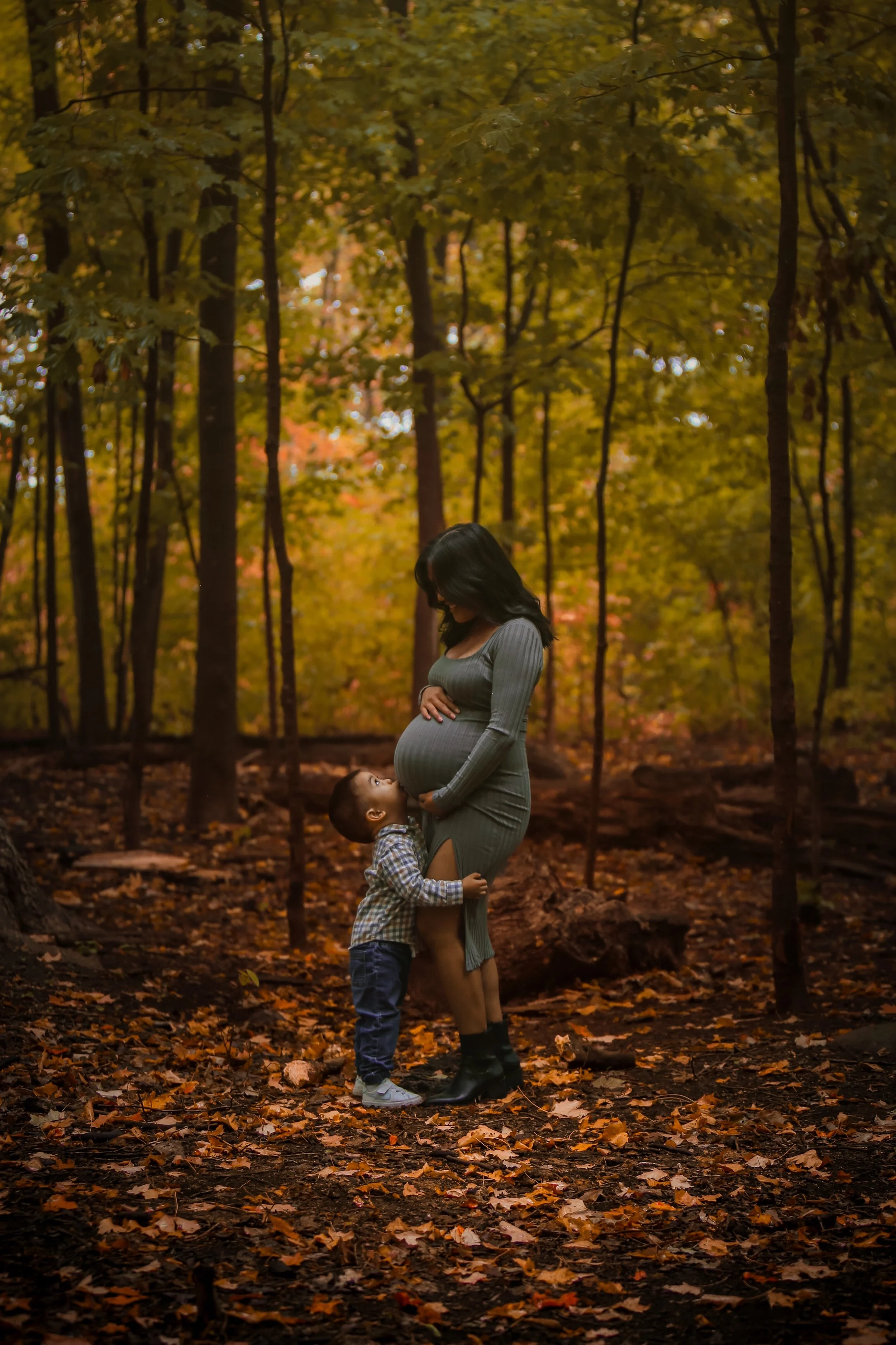 A pregnant woman in a gray dress and black boots standing in a forest with autumn leaves on the ground, while a young boy in a plaid shirt and jeans looks up at her and touches her belly.