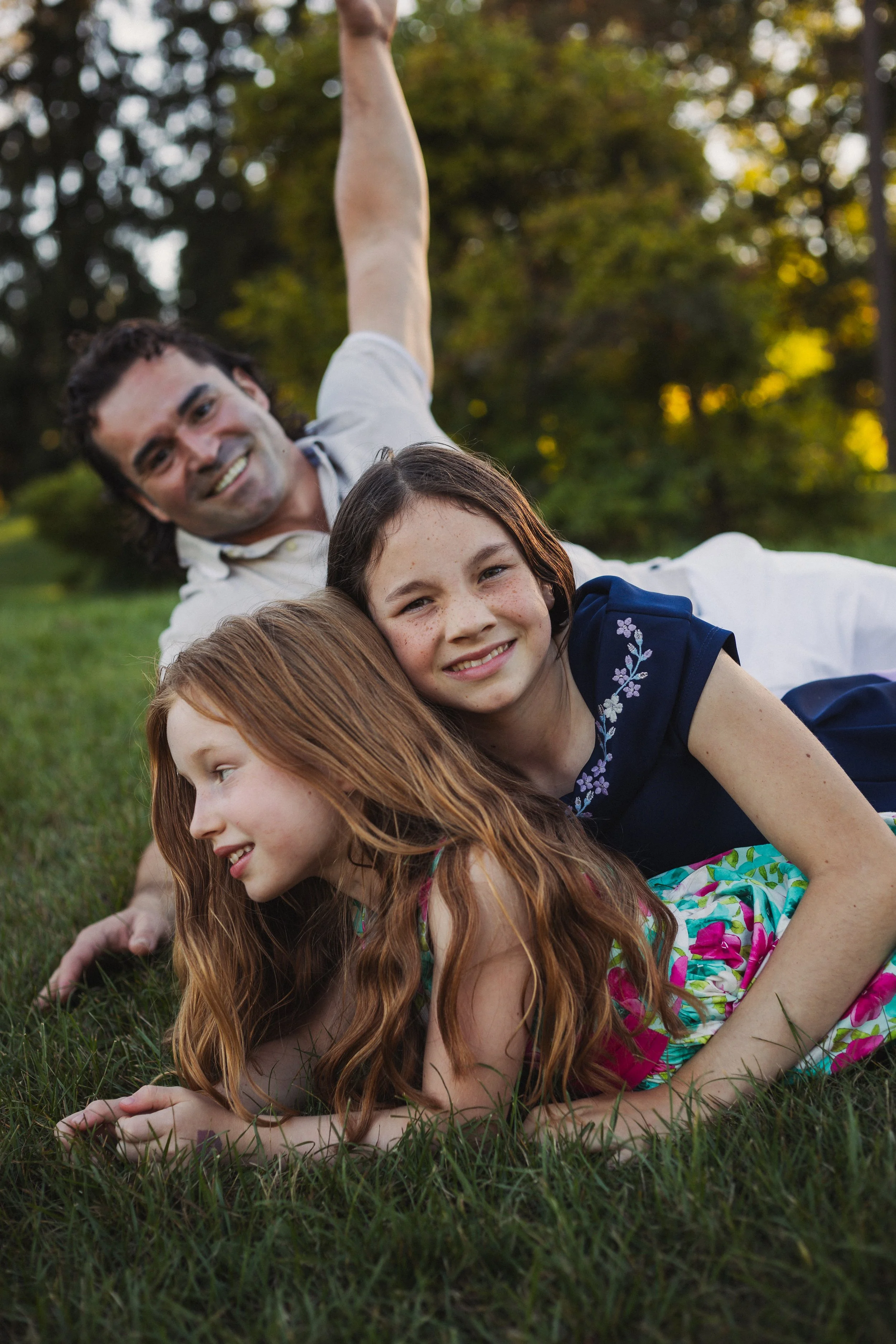 A family of three lying on the grass outdoors, with trees in the background. The father is smiling and lying on his side, the girl with brown hair and freckles is in the middle, and a red-haired girl in a colorful dress is on her side in the foreground.