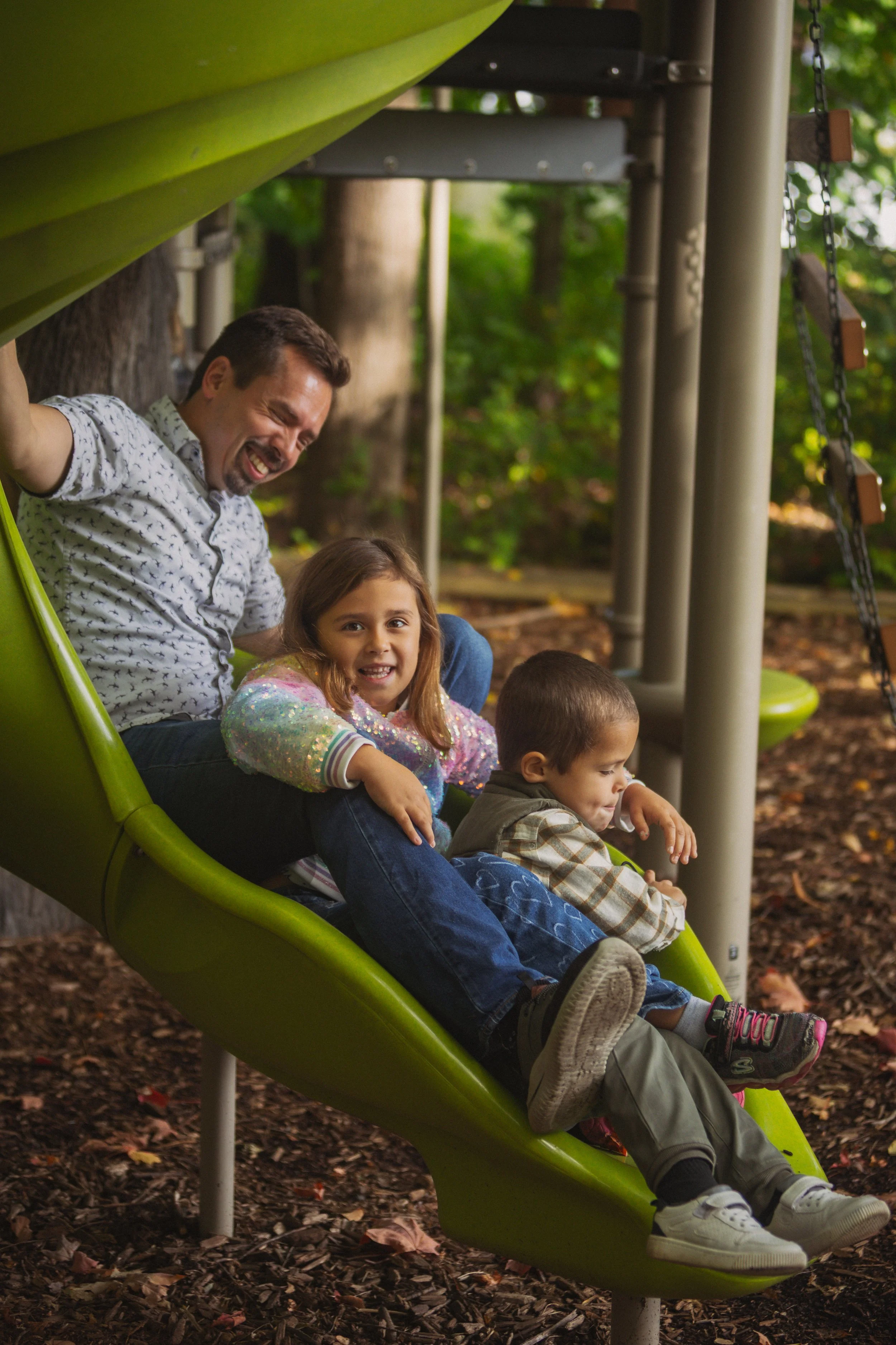 A man and two children sliding down a playground slide outdoors, surrounded by trees.